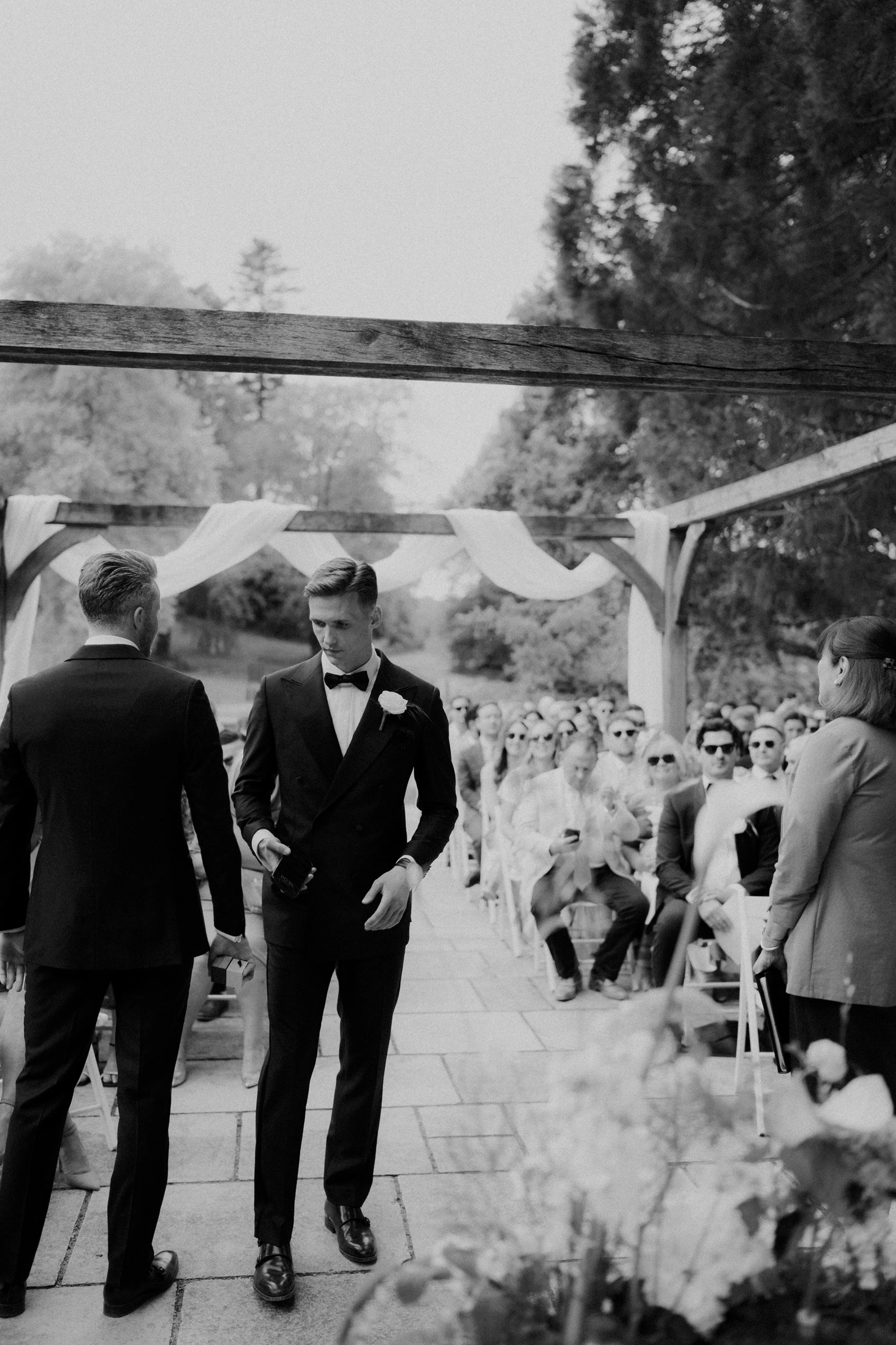 Black and white photo of a wedding ceremony outdoors, with two men in tuxedos at the altar and a seated audience in the background.