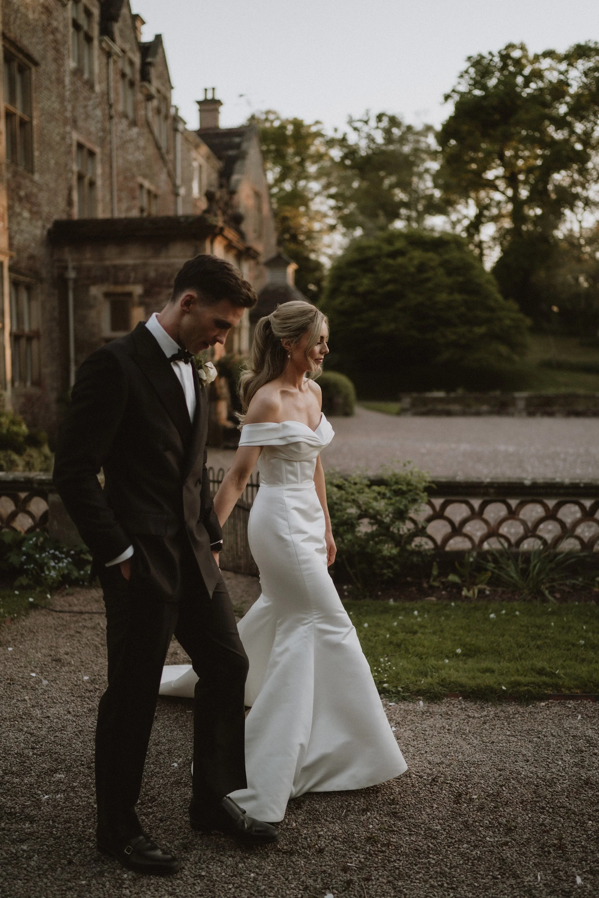 A bride and groom walking outdoors near a historic building during sunset, dressed in wedding attire, with the groom in a black tuxedo and the bride in a white off-the-shoulder wedding dress.