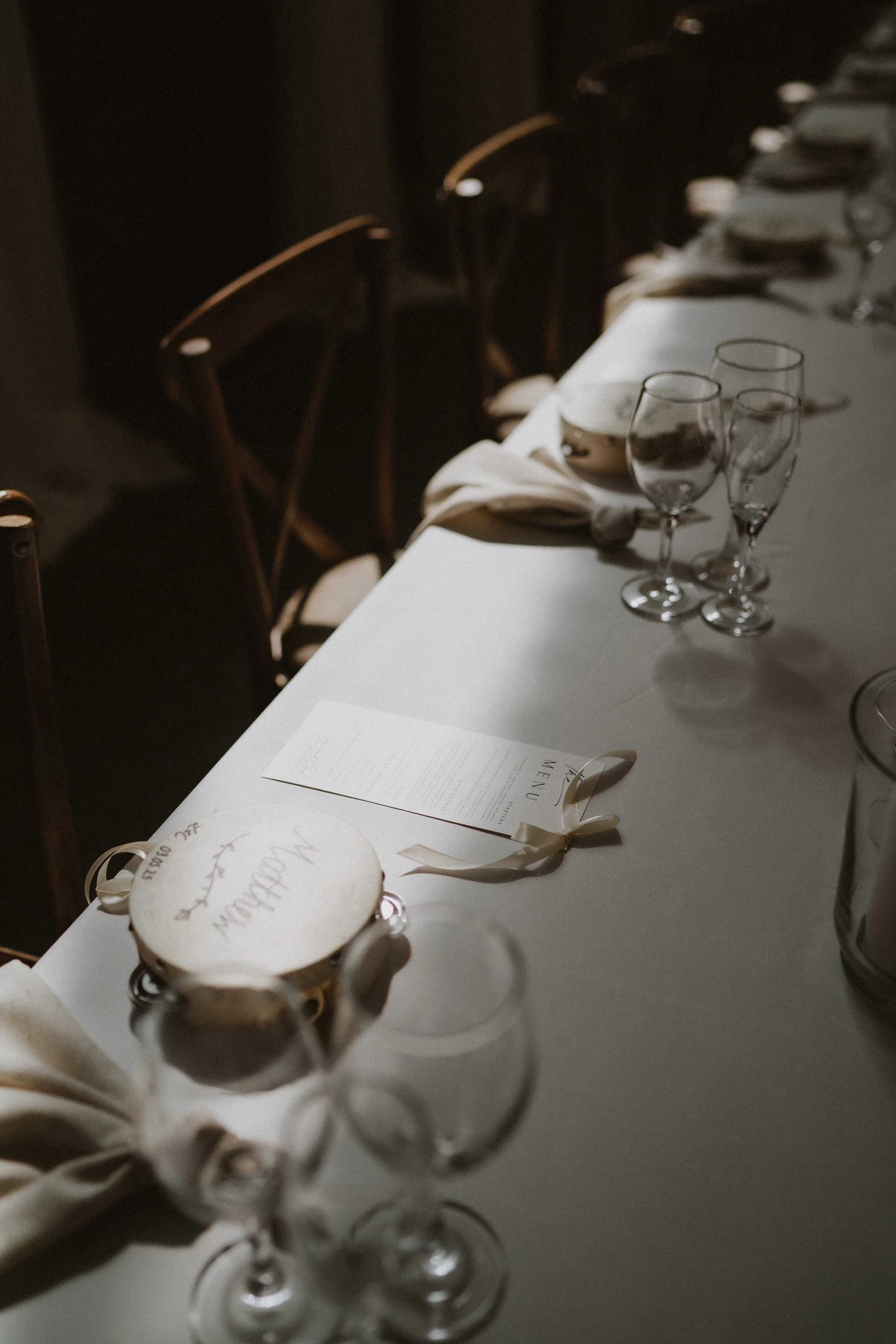 A formal dining table set with multiple empty wine glasses, a white tablecloth, napkins, a menu, and a small round cake with the words 'Wedding Day' written on it, in a dimly lit room.