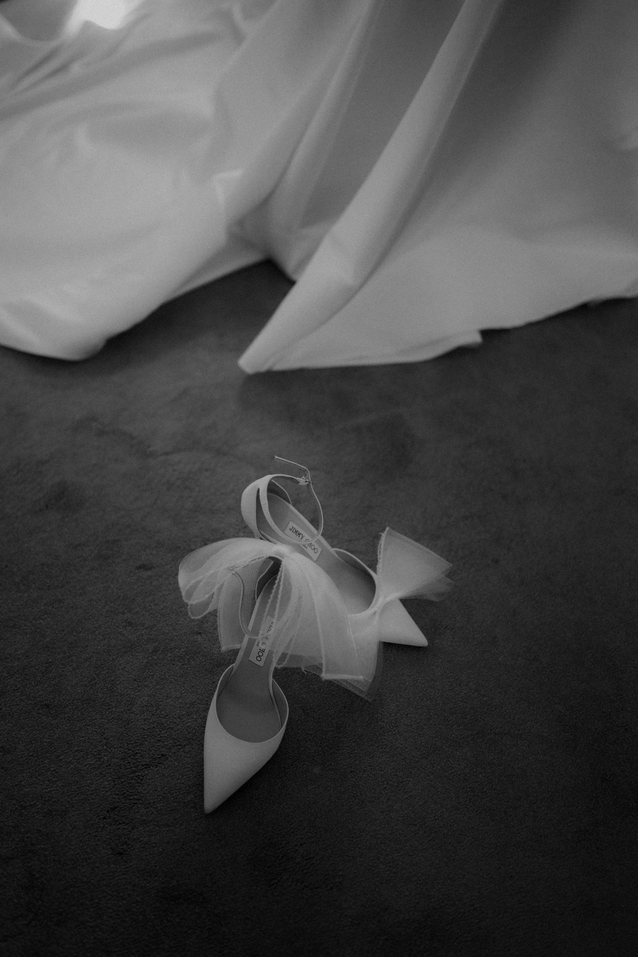 A single white high heel shoe with a tulle bow on the toe, lying on a carpeted floor near a bed or sofa with white bedding.