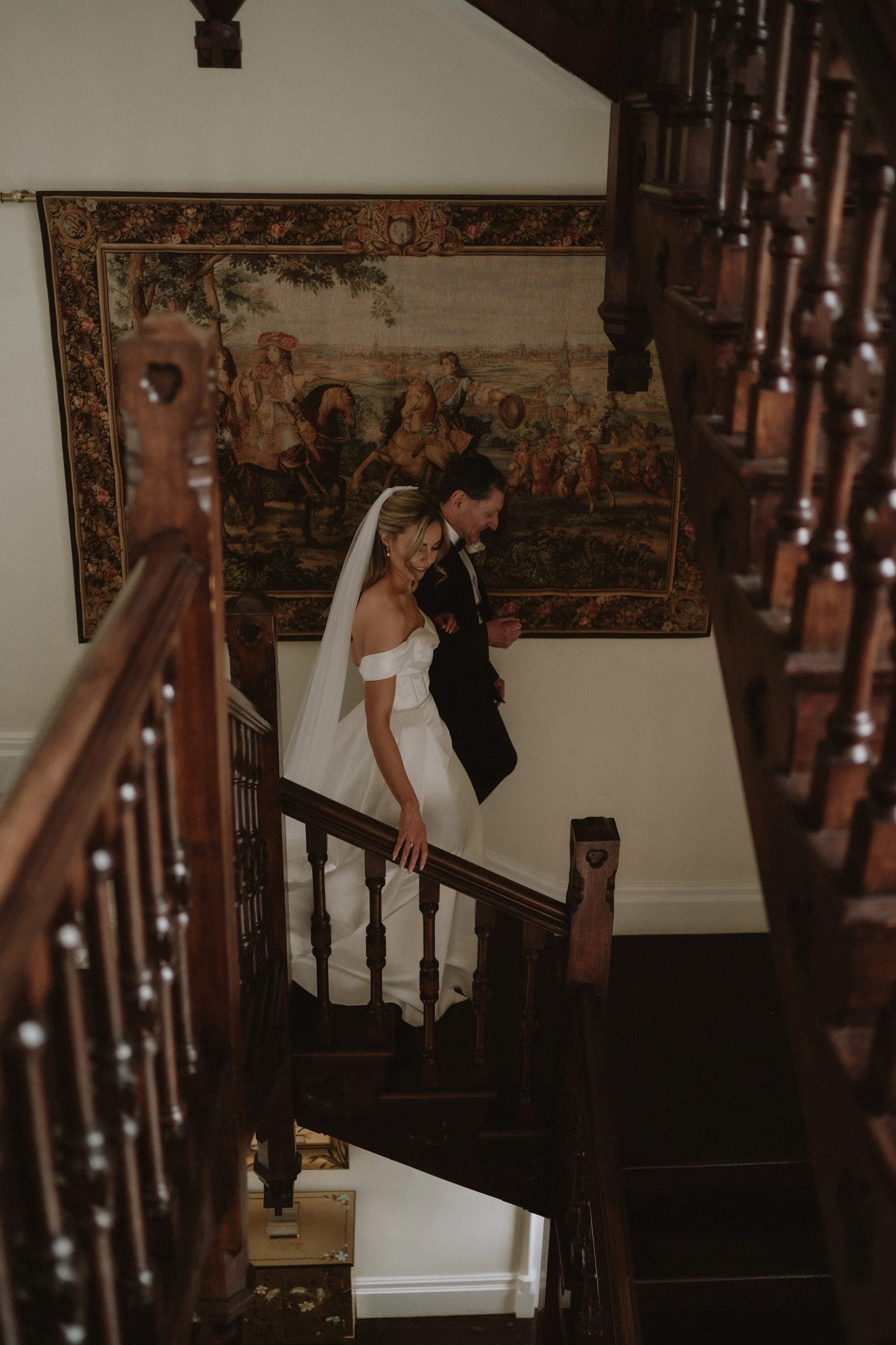 A bride in a white wedding dress and veil walking down a staircase with a groom in a black suit behind her, descending together.
