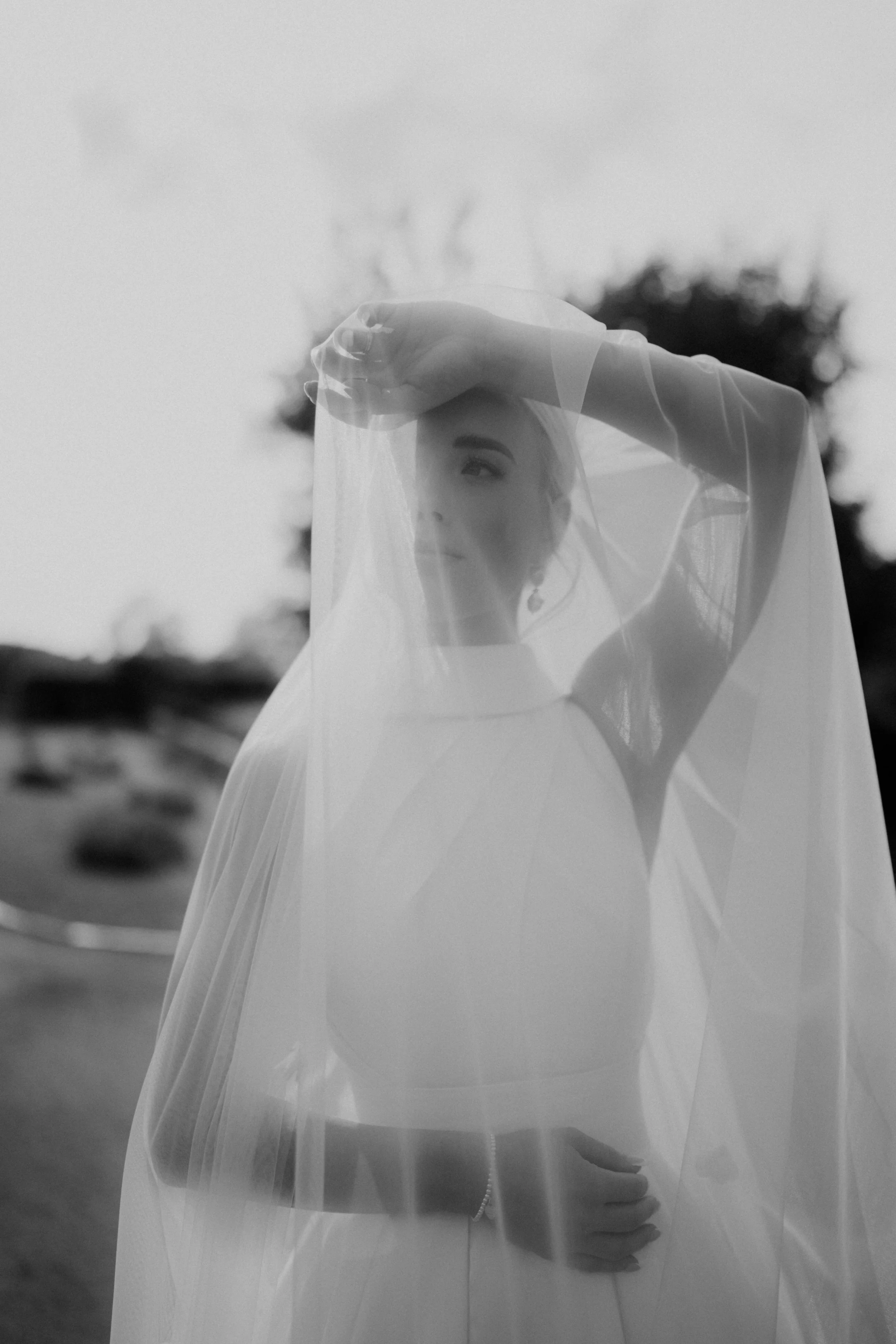 Black and white photo of a woman in a wedding dress standing outdoors, partially covered by a sheer veil. She has blonde hair styled up and is looking to the side with one arm raised above her head, resting on her forehead, with a blurred natural lan
