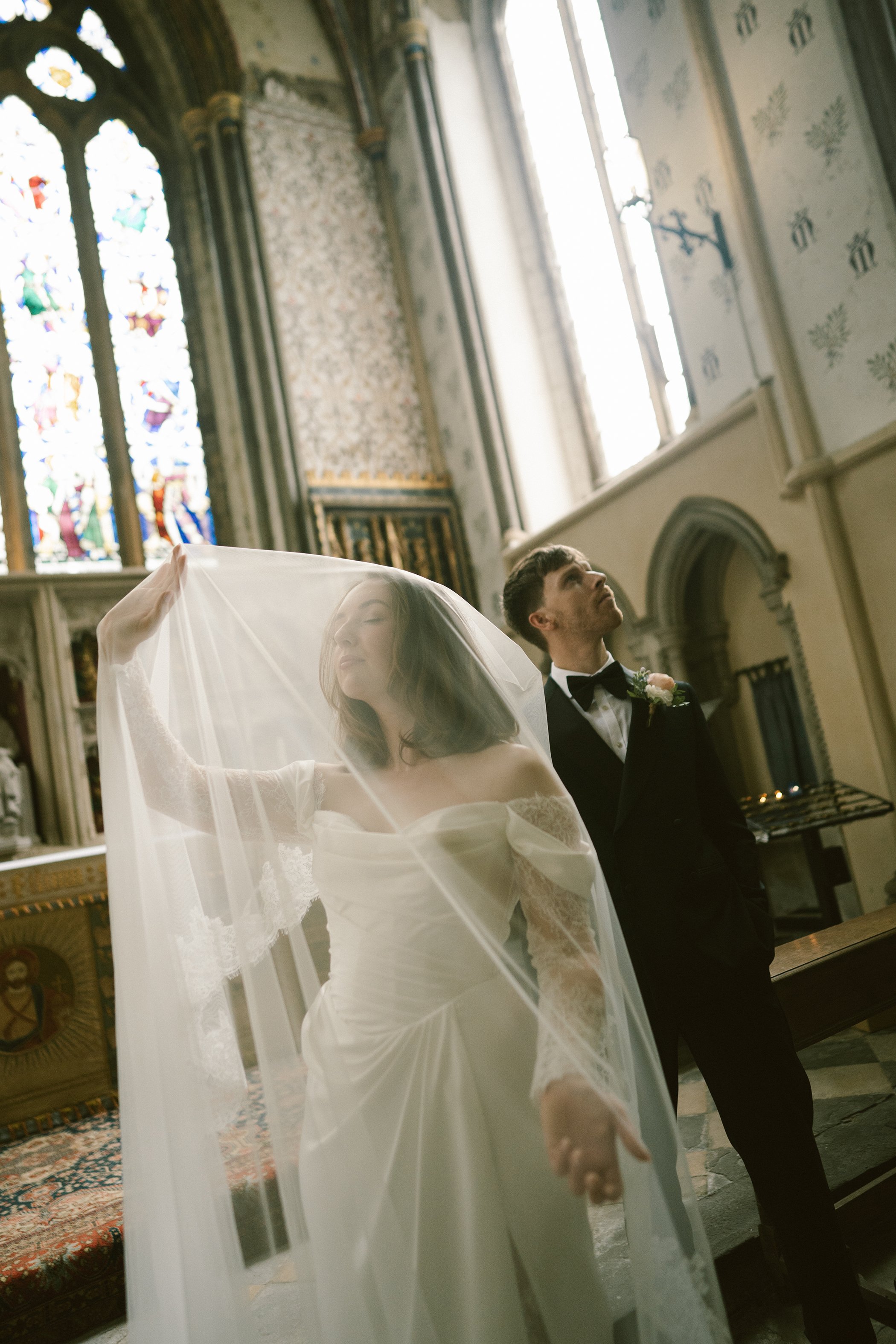 A bride and groom inside a church during their wedding ceremony. The bride has a white dress and veil, and the groom is in a black tuxedo with a bow tie. They are standing before an altar with stained glass windows in the background.