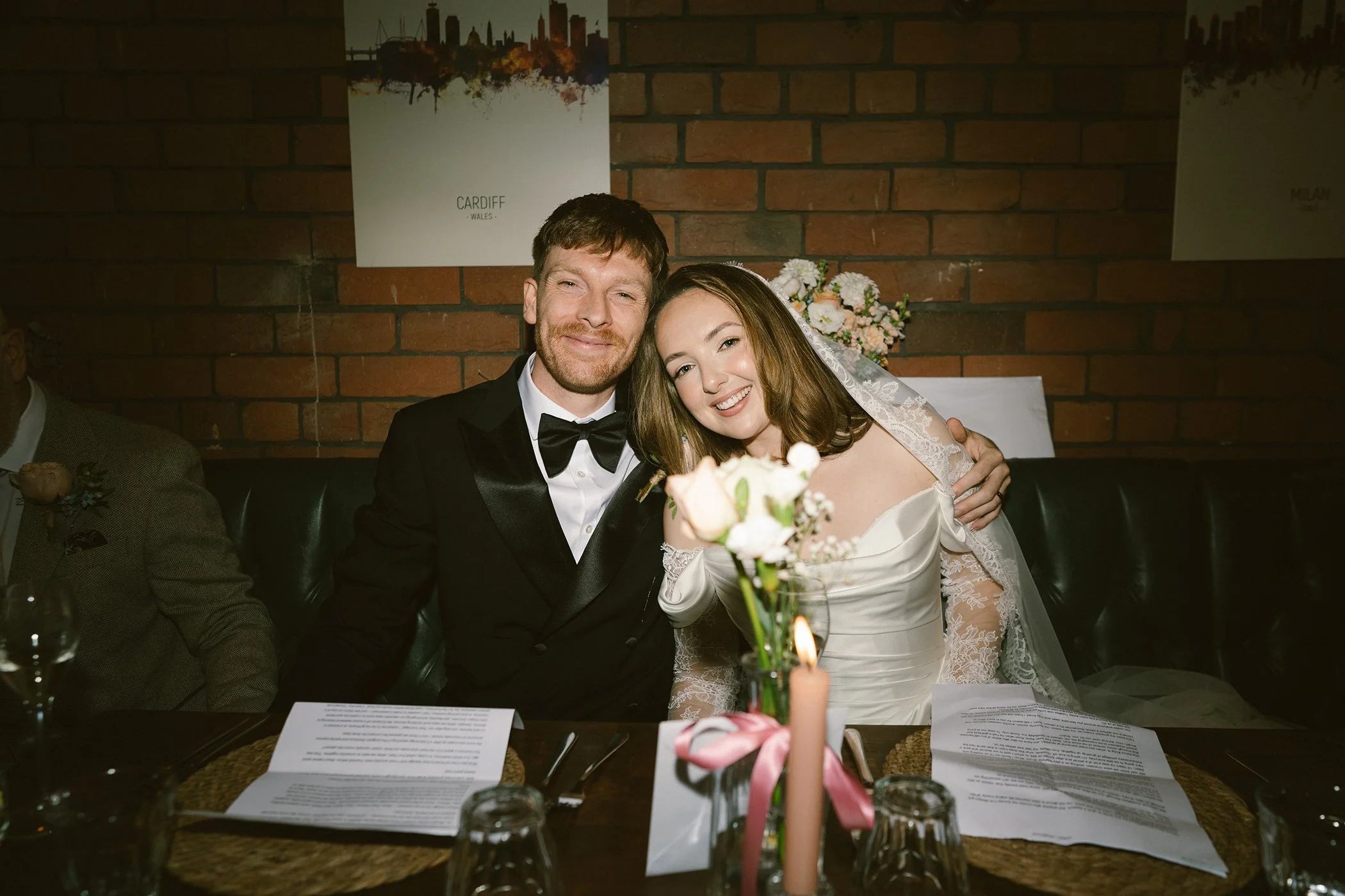 A bride and groom smiling and sitting close together at their wedding reception, with a brick wall and cityscape poster in the background. The bride wears a lace wedding dress and veil, holding a bouquet of flowers. The groom wears a black tuxedo wit
