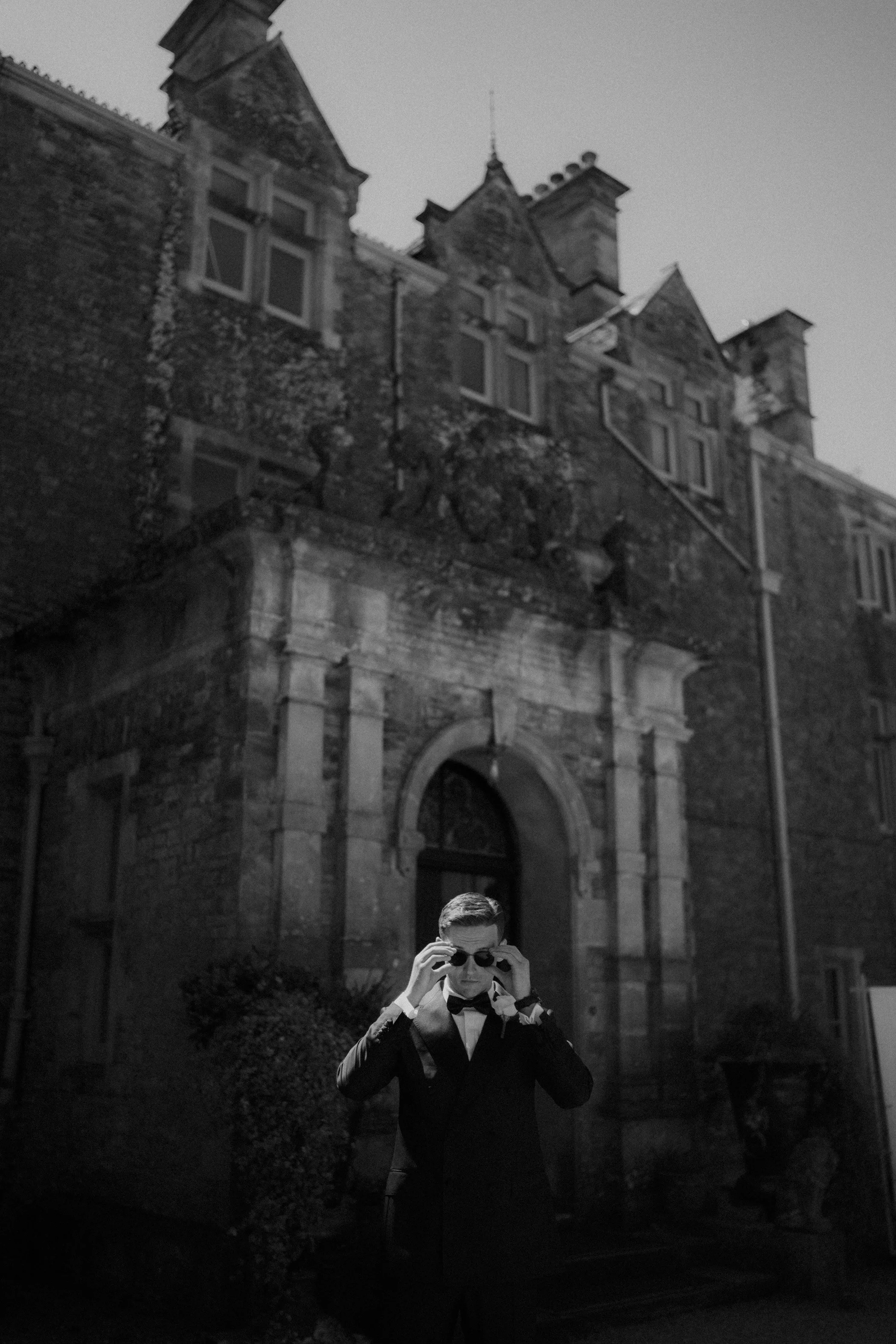 A man dressed in a tuxedo adjusts his sunglasses outside an old brick building.