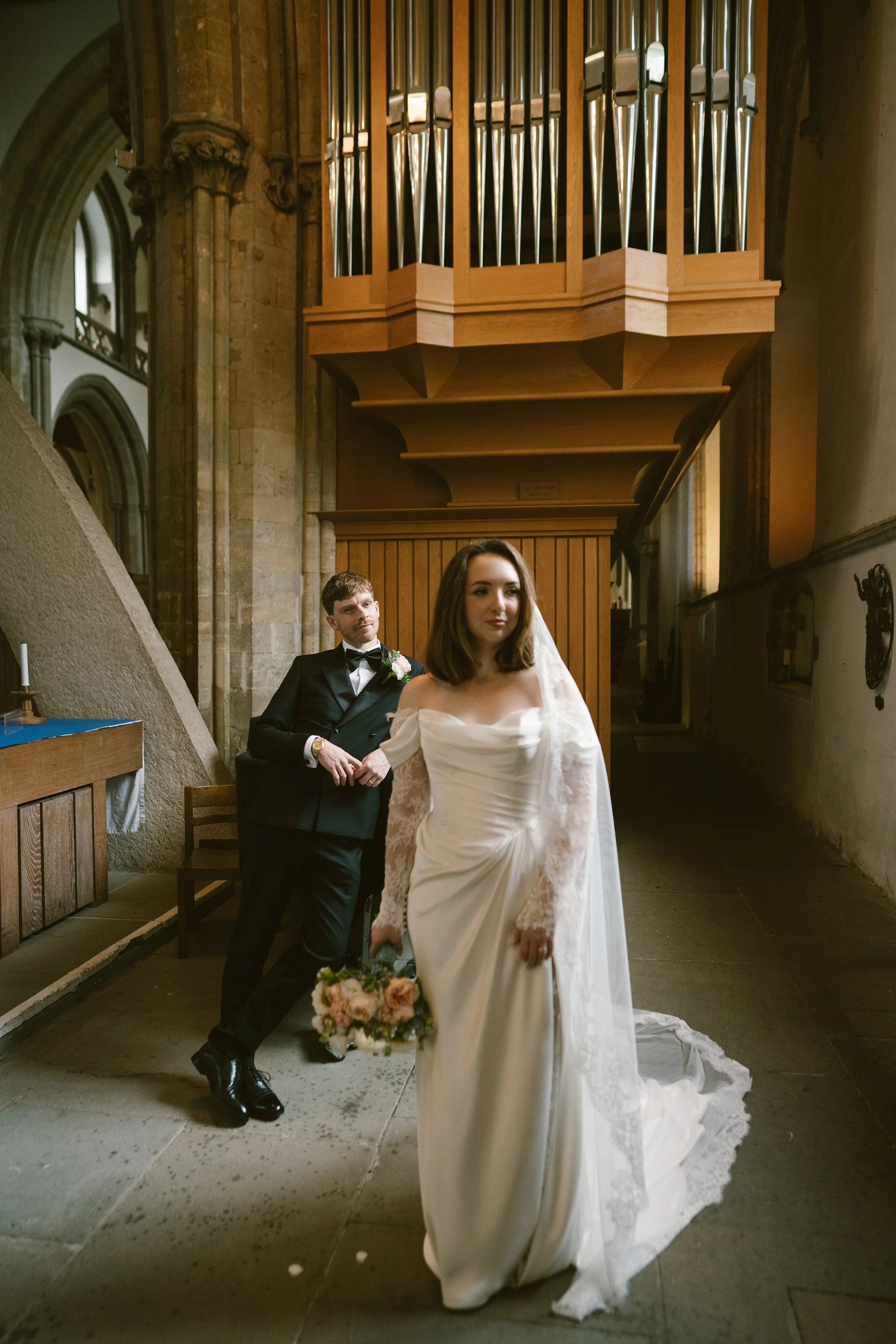 A bride in a white wedding dress walking down the aisle holding a bouquet of pink and white flowers, with a groom in a black suit and bow tie sitting in the background inside a church.