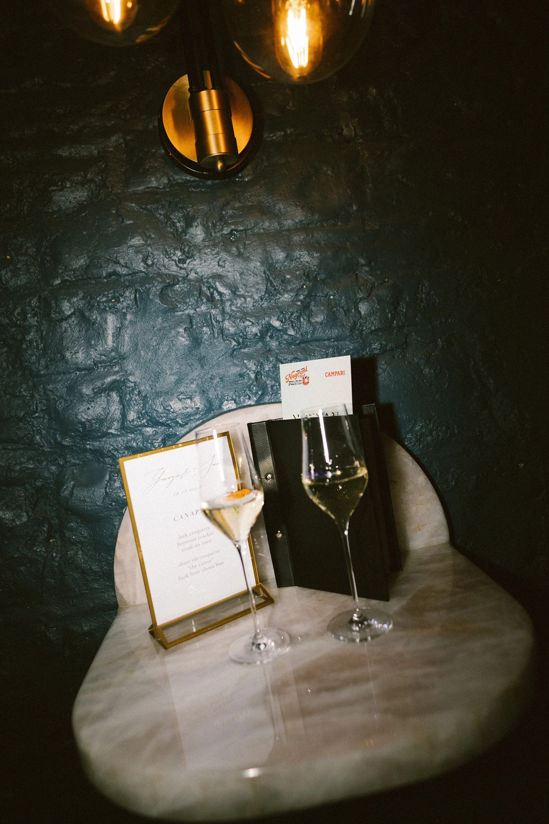 Two glasses of white wine on a marble table, with a menu and a black box holding a card that reads 'Campari' in the background. The scene is set against a dark textured wall with warm lighting.