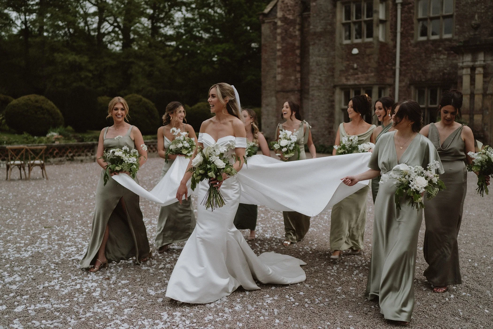 Bride in a white wedding gown and veil walking with her bridesmaids in olive green dresses, outdoors at a historic stone building, holding bouquets, on a gravel path with flower petals.