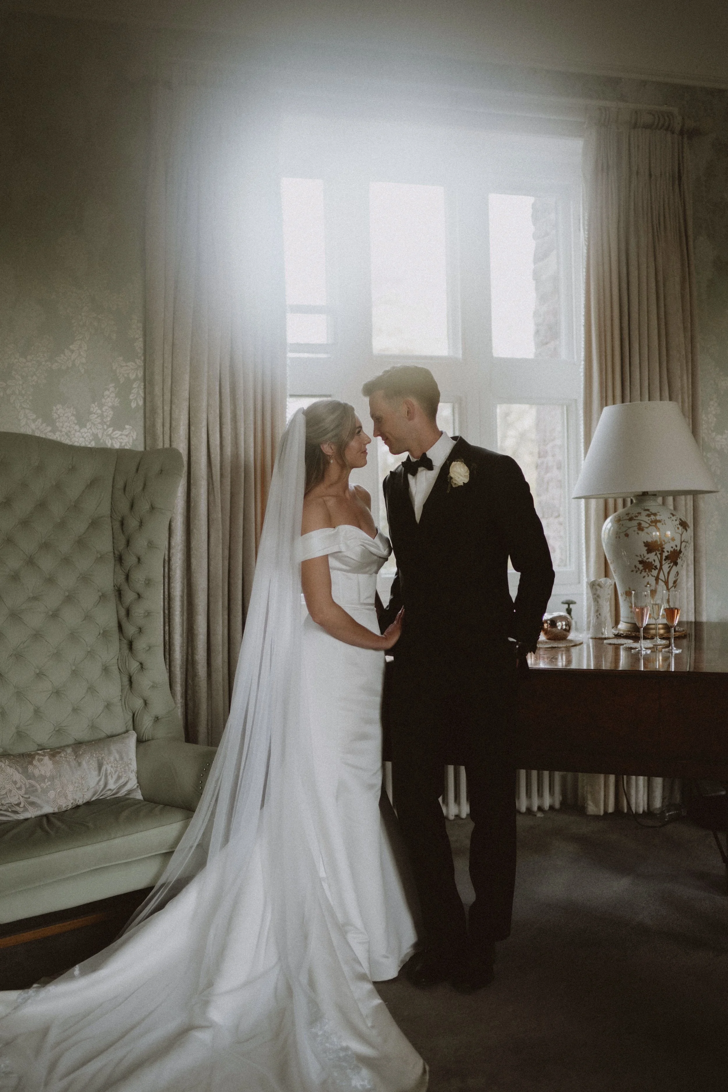 A bride in a white wedding dress and veil is standing close to a groom in a black tuxedo, inside a warmly lit room with a large window and decorative curtains.