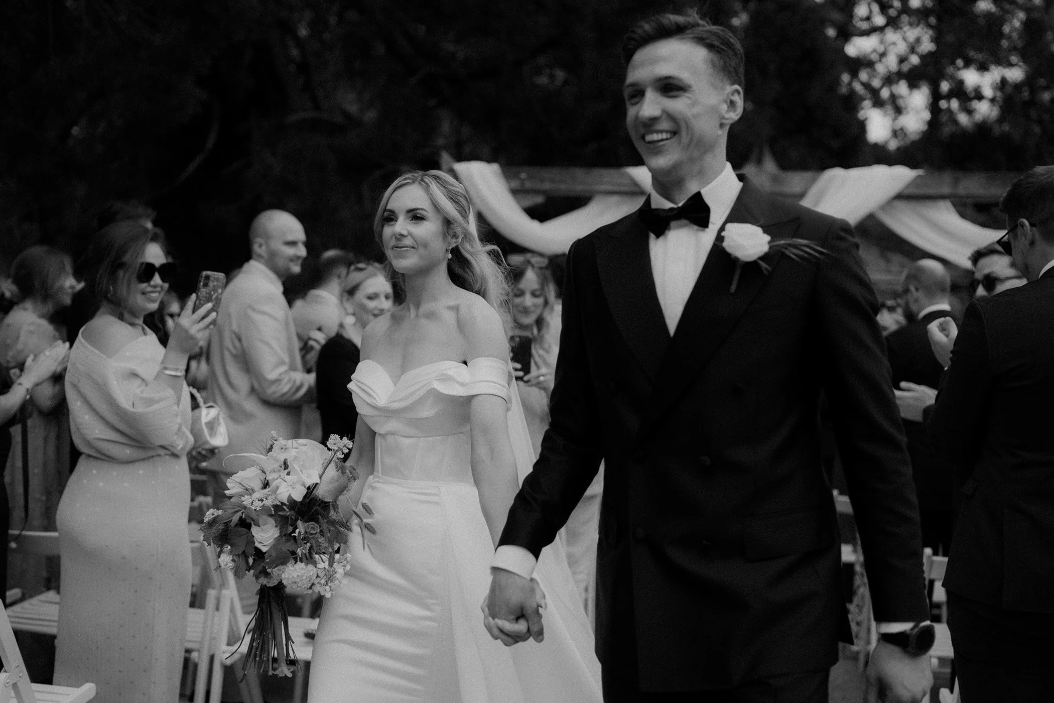 Black and white photo of a wedding ceremony showing a smiling bride holding a bouquet and a groom holding her hand, with guests in the background clapping and taking photos outdoors.