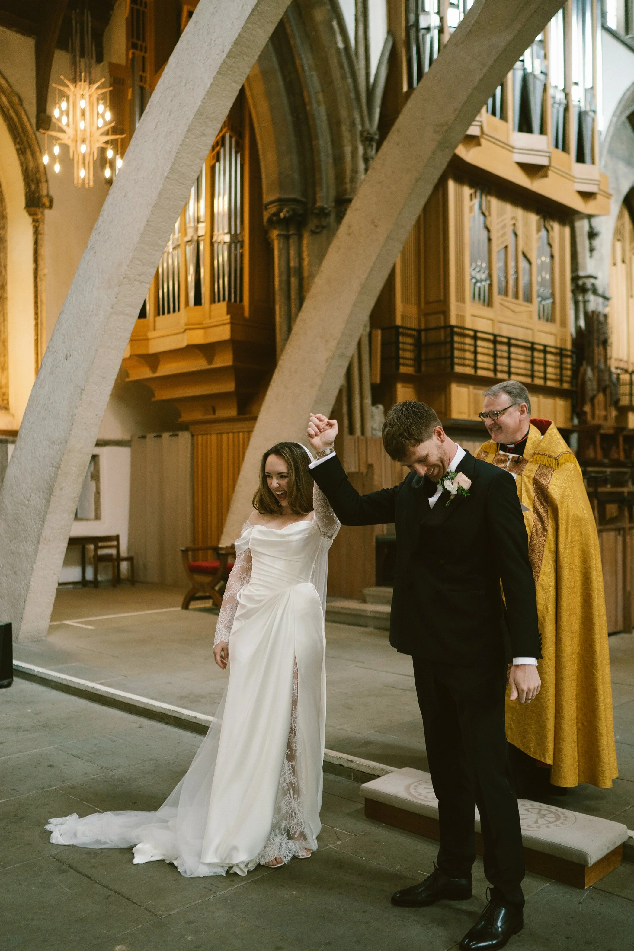 A wedding ceremony inside a church with a bride and groom holding hands and smiling at each other, with a priest in a golden robe officiating.