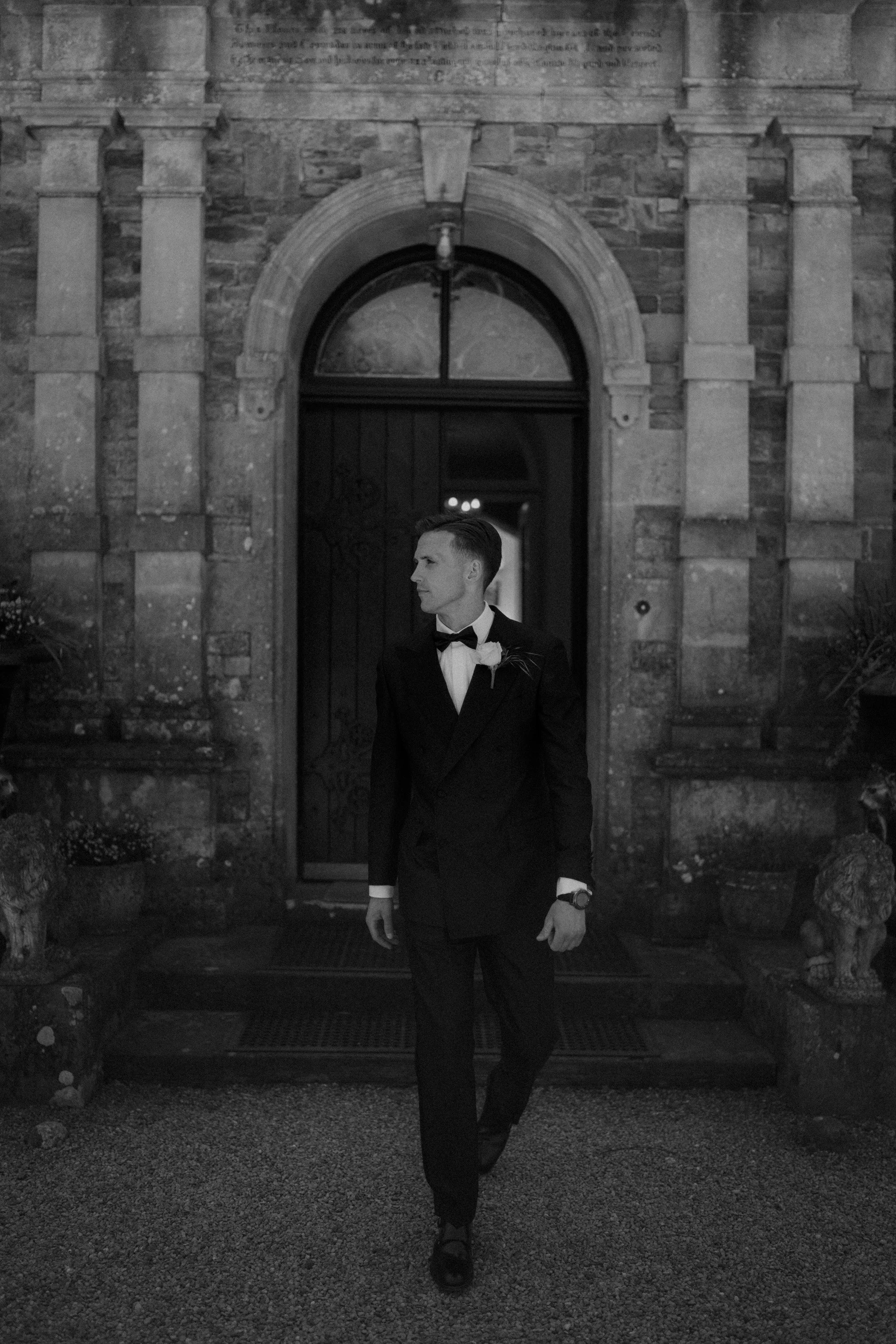 A man in a tuxedo, holding a boutonniere, walking out of a stone building with an arched door at night.