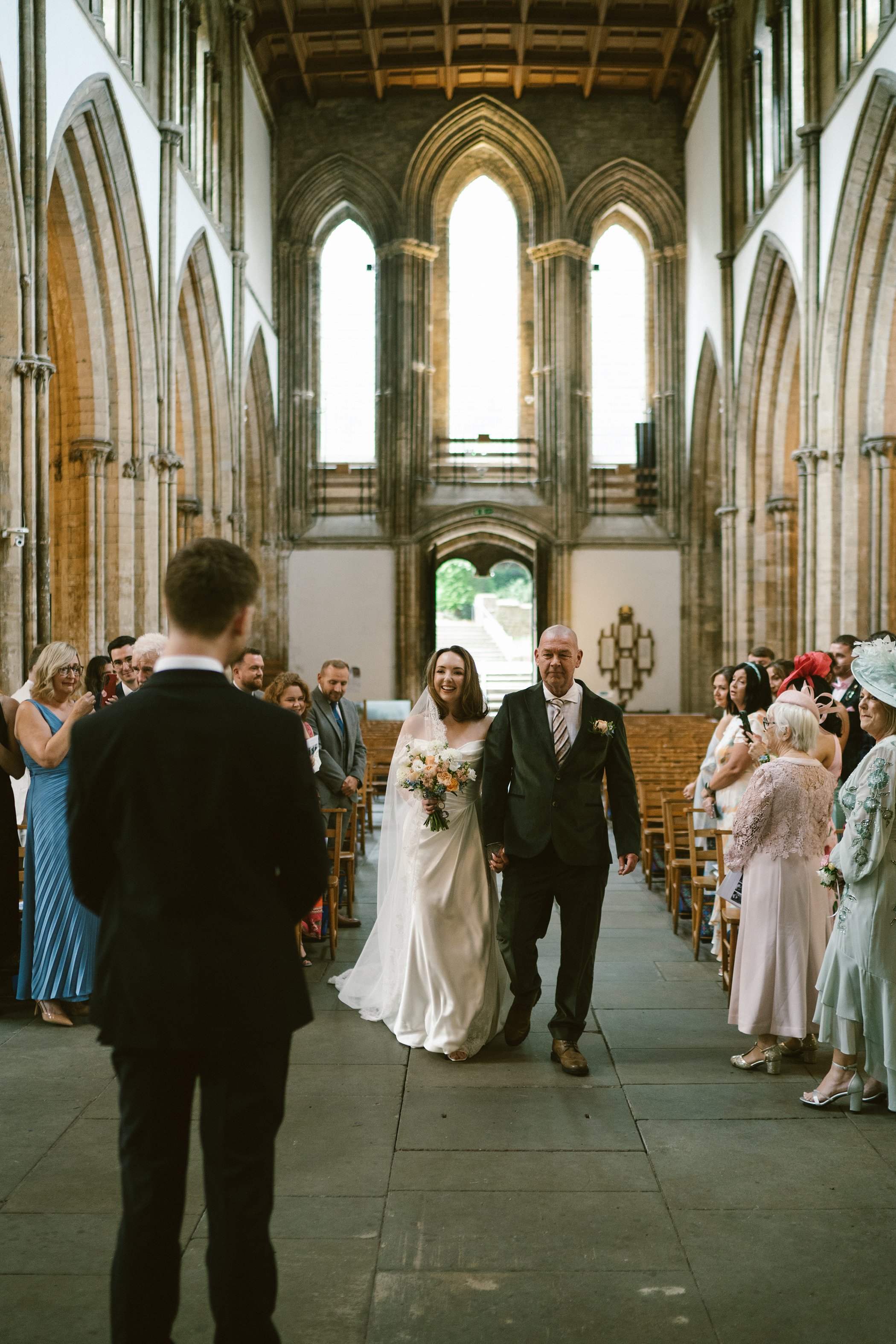 A bride walking down the aisle holding a bouquet, accompanied by an older man, possibly her father, during a wedding ceremony in a church with tall stained-glass windows and Gothic architecture. Guests are seated on either side, smiling and watching.