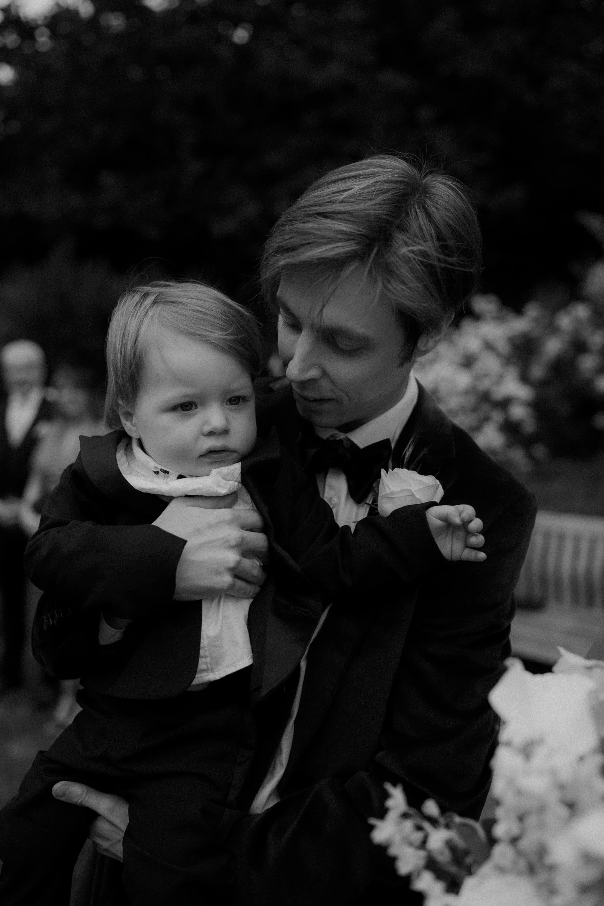 A man in a tuxedo holding a young child, both dressed formally, outdoors surrounded by blurred people and flowers.