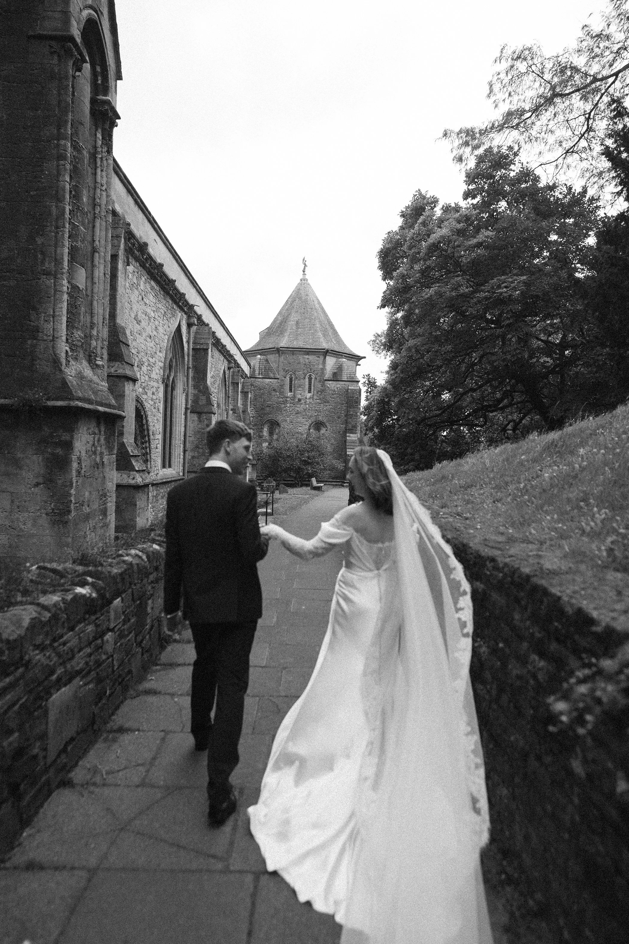 A bride and groom walking outdoors on a paved path, with a historic stone building and trees in the background. The bride is wearing a long white wedding gown, and the groom is dressed in a black suit. The couple is holding hands, and the bride's gow