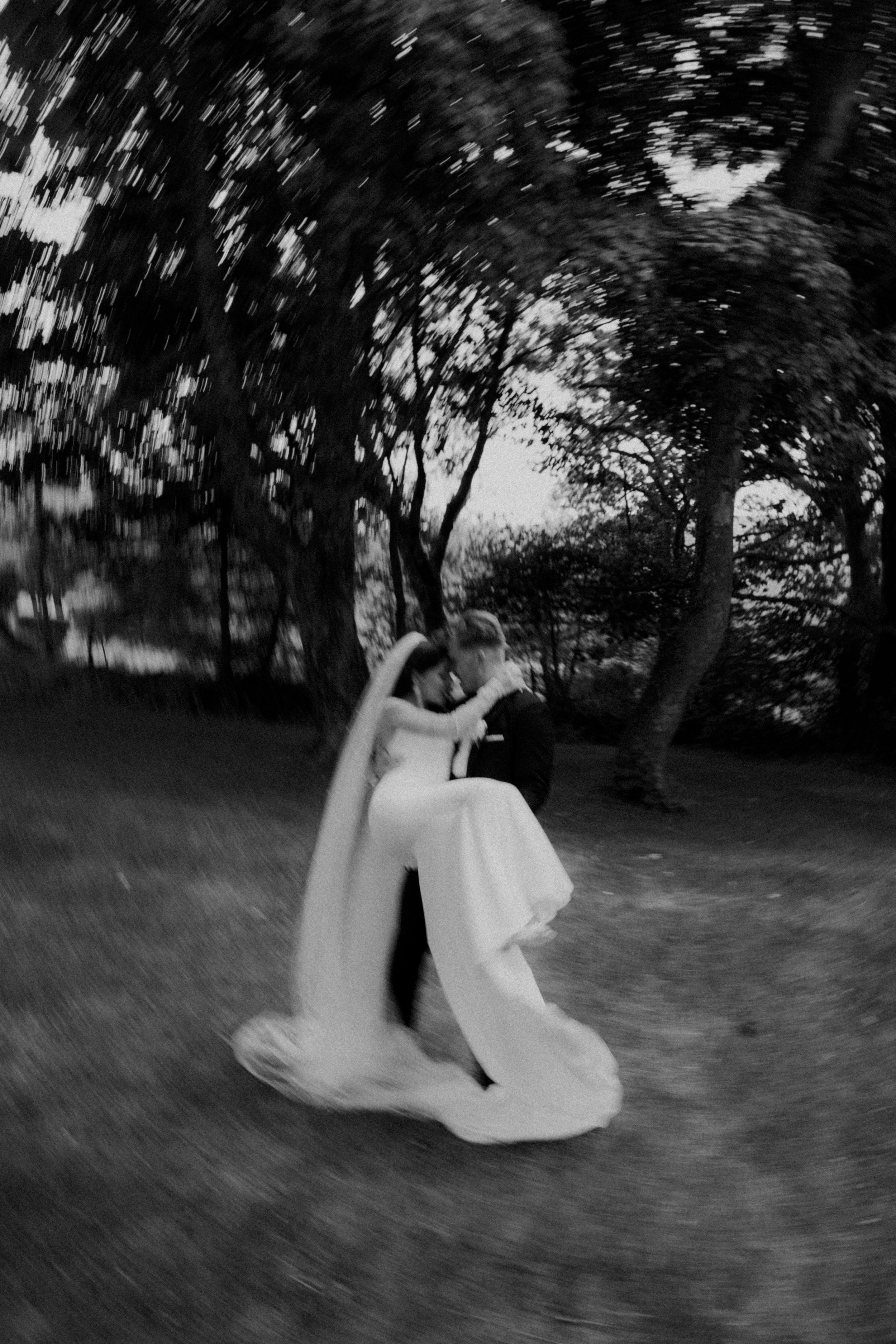 Black and white photo of a bride and groom dancing outdoors among trees, with motion blur creating a swirling effect.