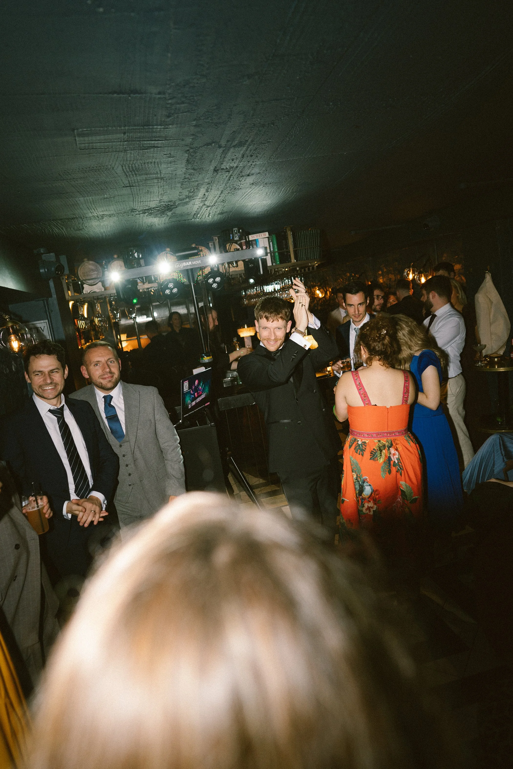 People dancing and socializing in a dimly lit bar or club with a DJ booth in the background.