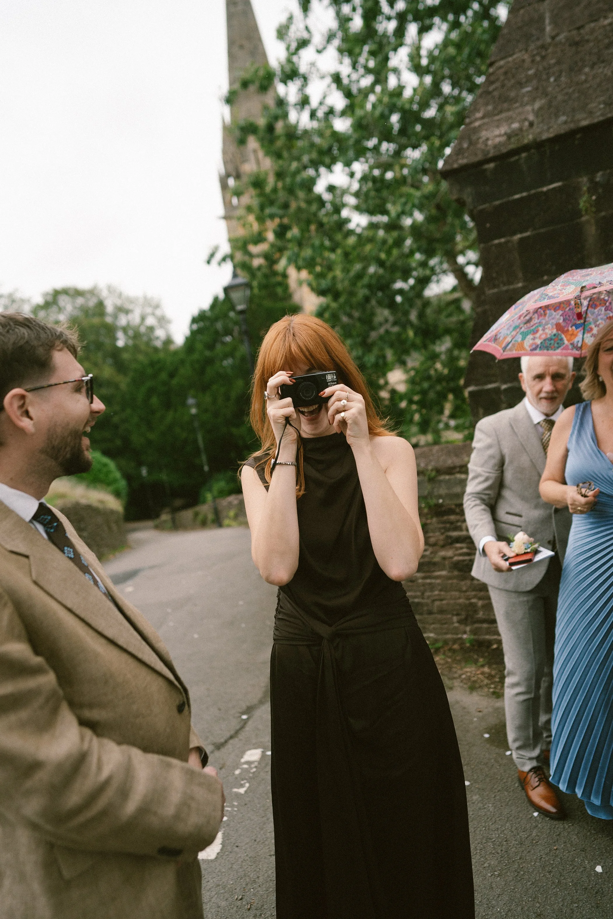 A group of people standing outdoors, with a woman with red hair taking a picture with a camera, and others around her dressed in formal attire, some holding umbrellas.