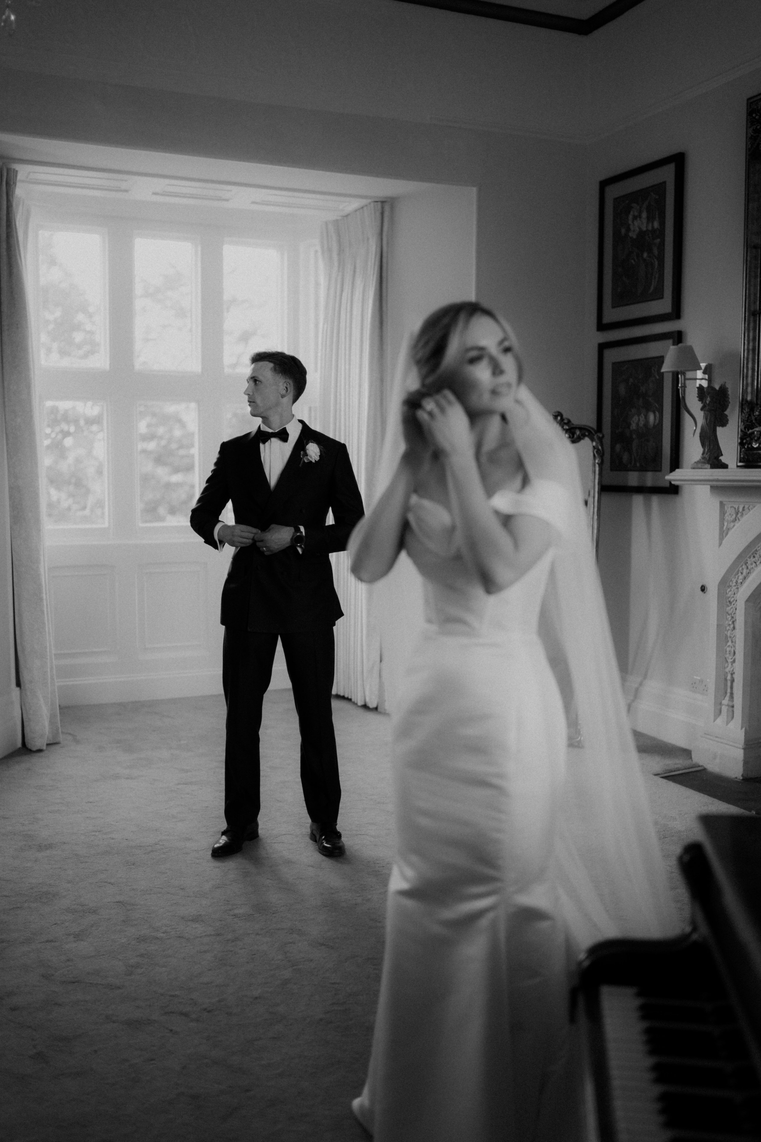 A black and white photo of a bride adjusting her hair and a groom in a tuxedo looking out the window in a decorated room.