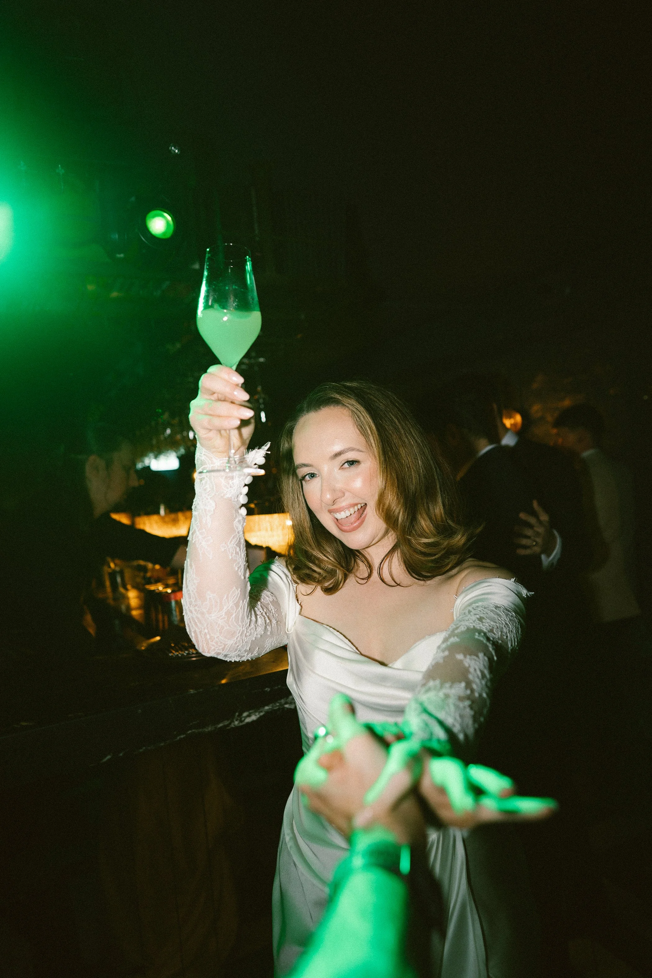 A woman in a wedding dress smiling and holding a champagne flute, with a green laser beam reaching out from her hand, at a dimly lit party or celebration.