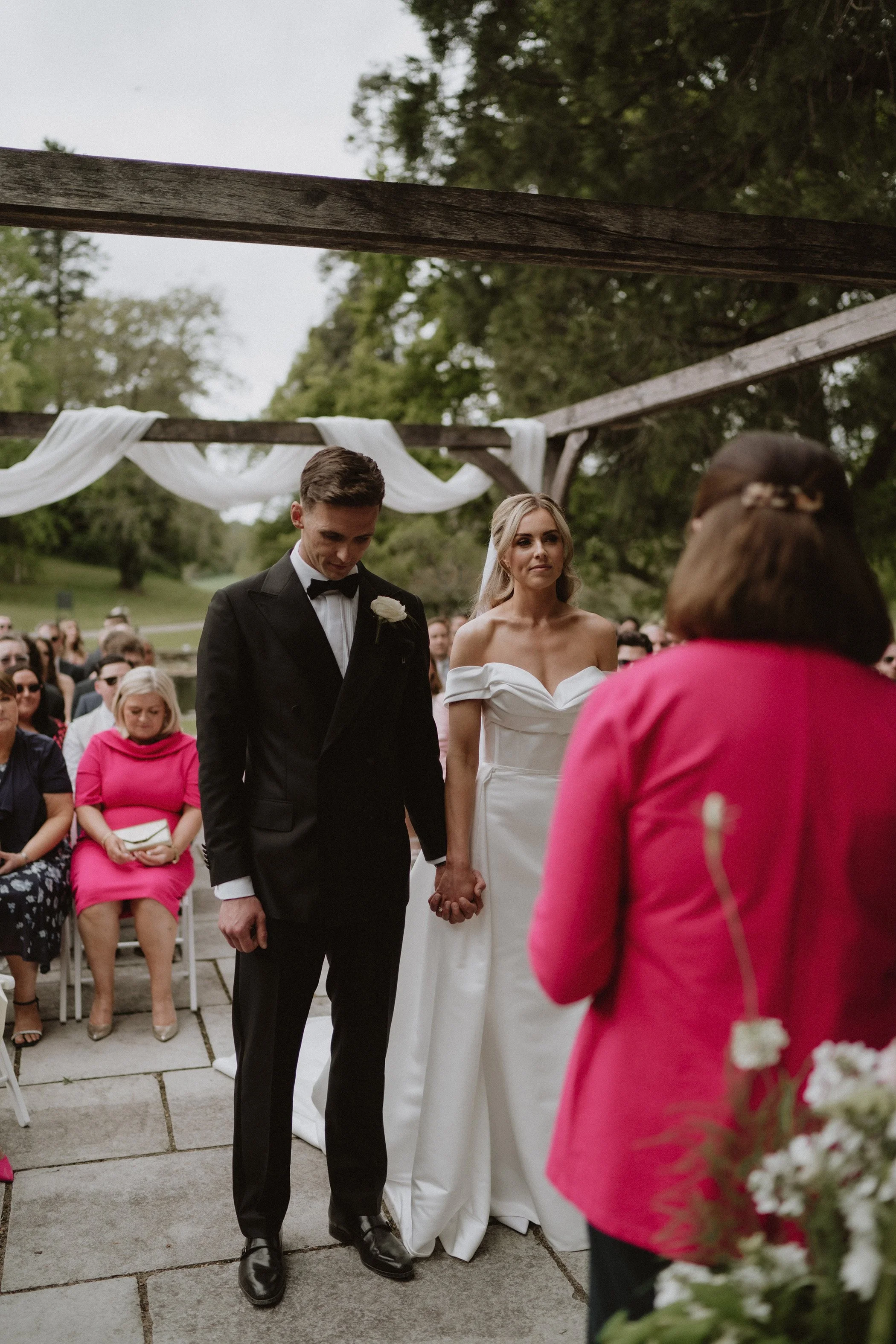 A bride and groom holding hands during their outdoor wedding ceremony. The groom is dressed in a black tuxedo, and the bride is wearing a white off-the-shoulder gown. Guests are seated in the background, under a wooden arch decorated with white drape