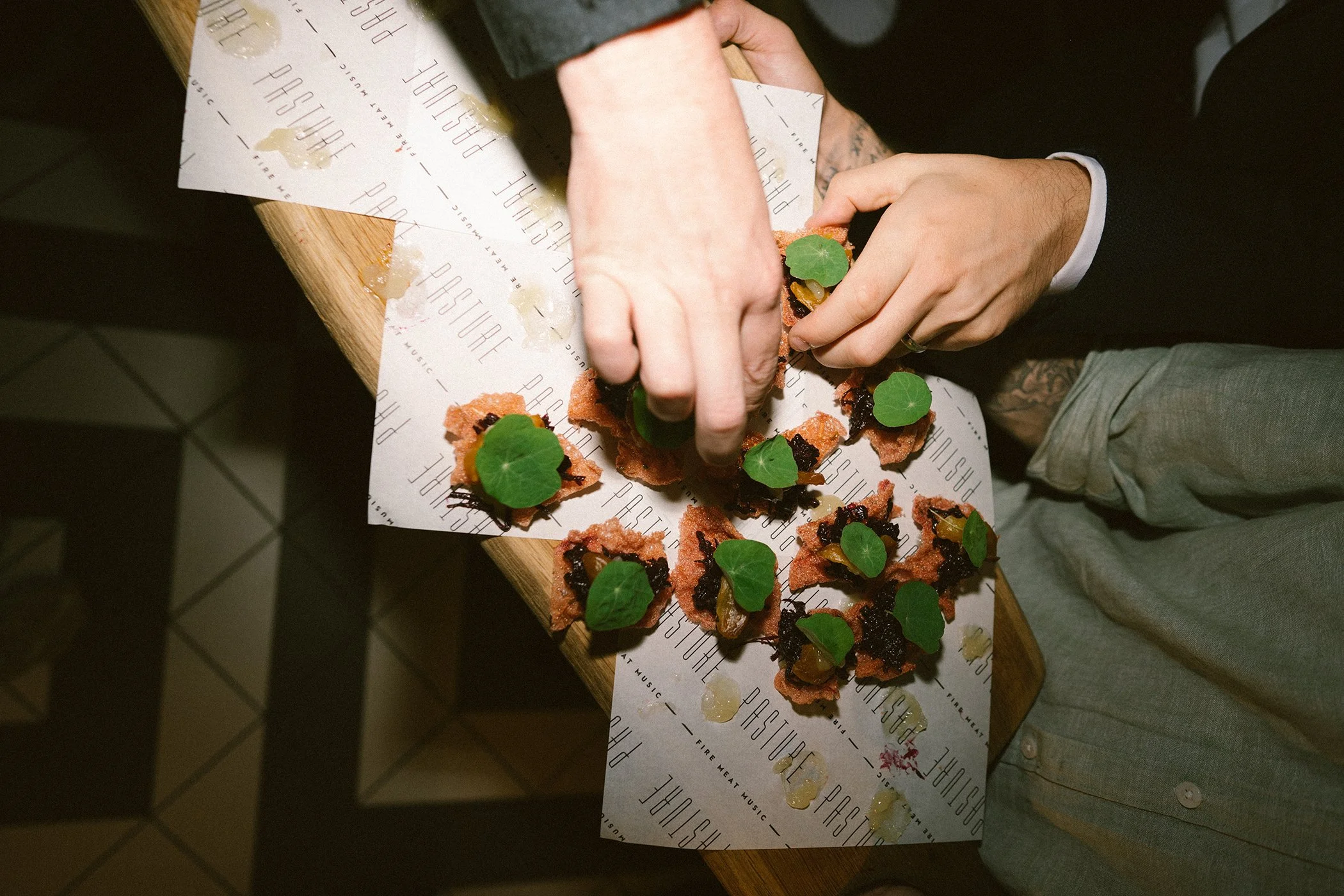 Person preparing or serving a dish with bread, herbs, greens, and dark sauce on a paper-lined wooden surface.