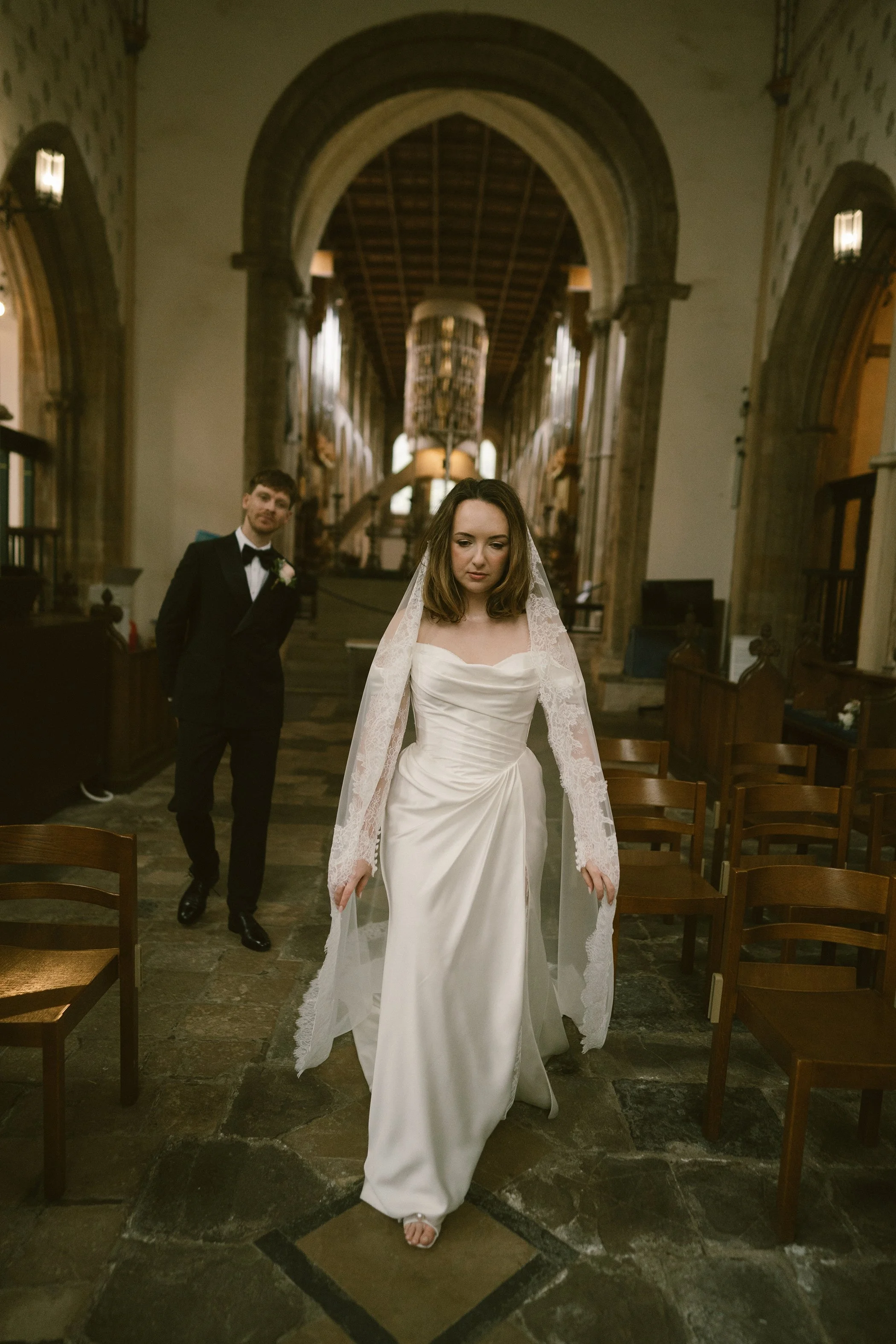 A woman in a white wedding dress with lace sleeves and a veil walks down the aisle of a church, with a man in a black tuxedo and bow tie standing in the background.