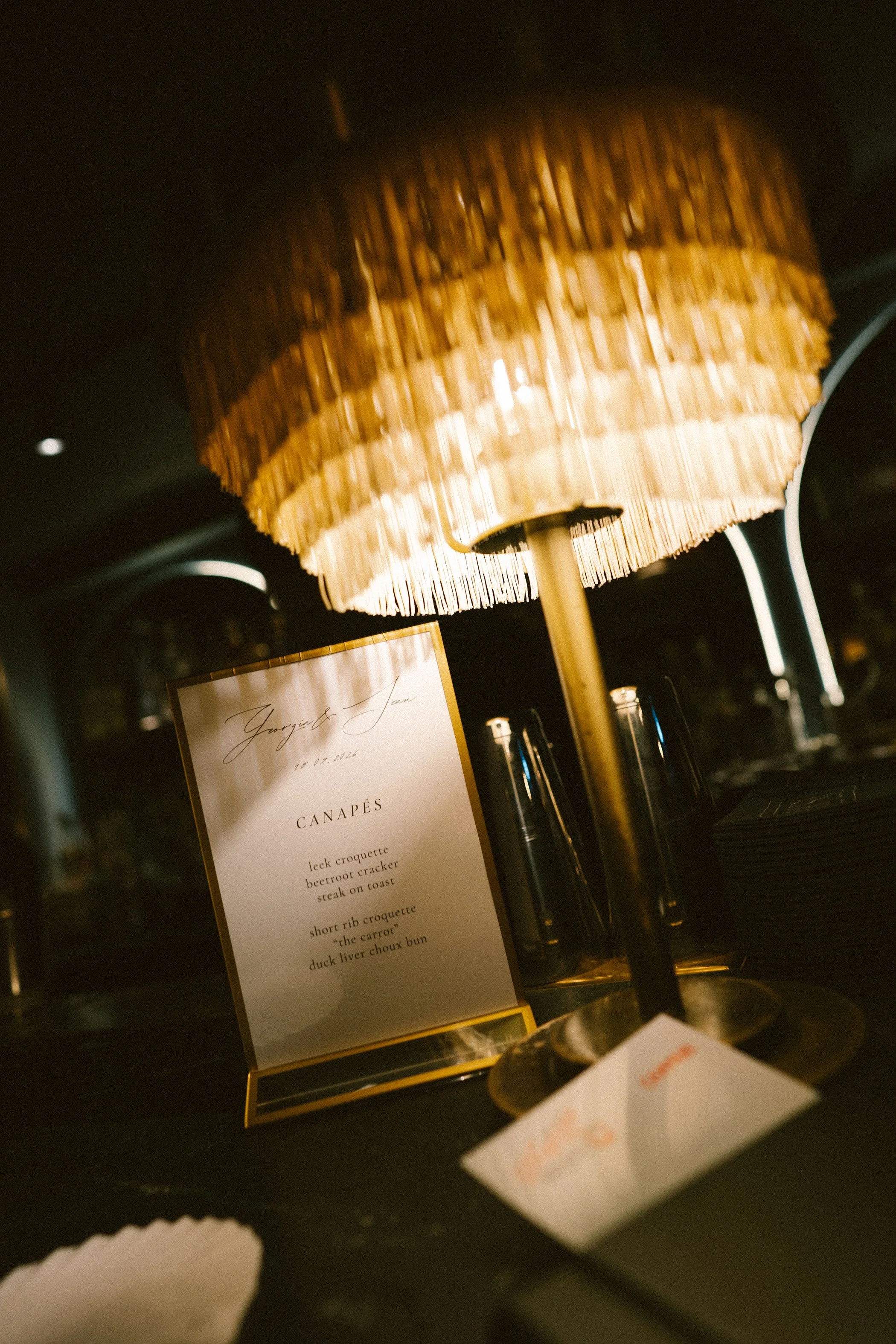 A table lamp with a woven, bamboo-style lampshade emitting warm light, positioned above a menu on a dark table surface with bottles and other objects in the background.