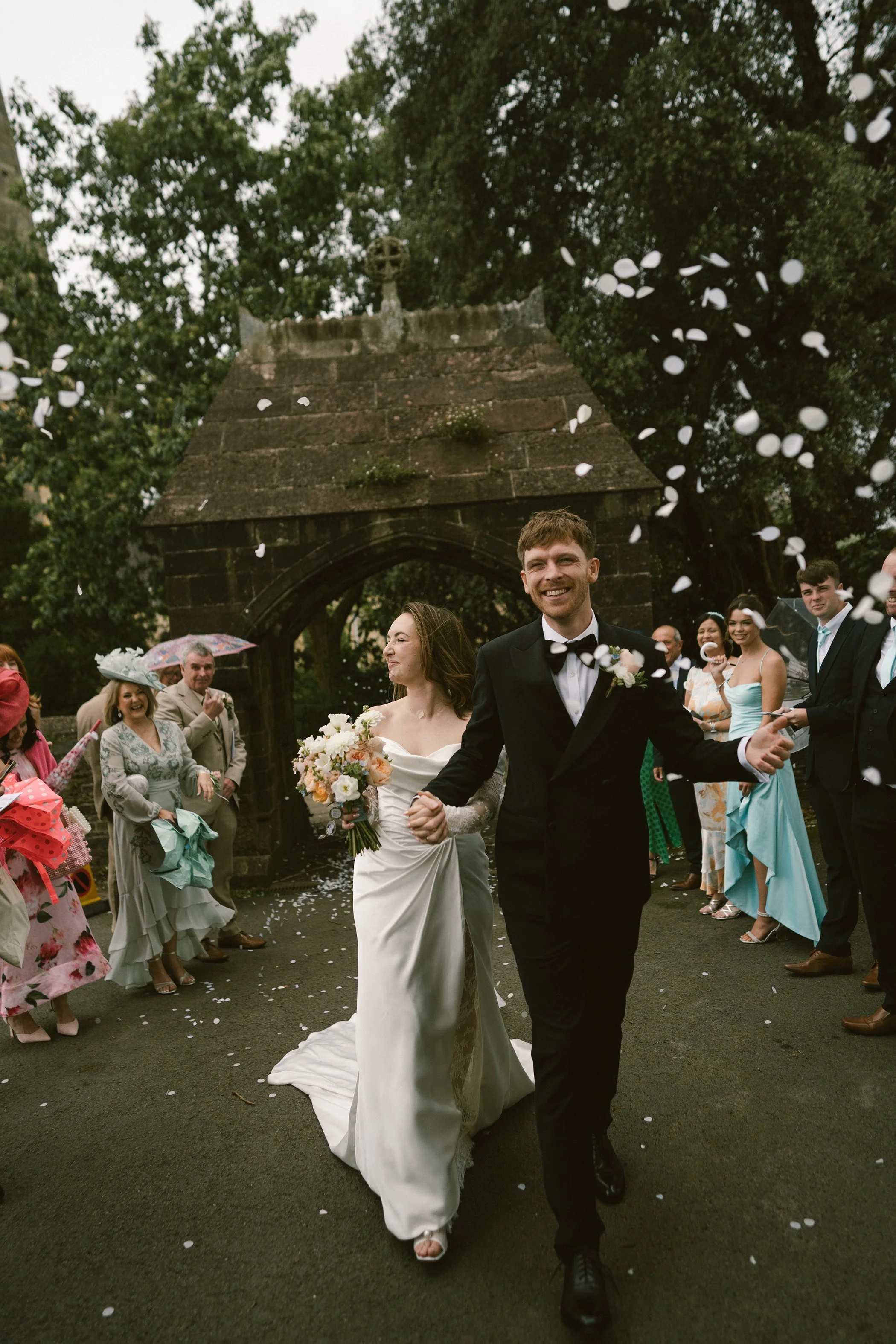 A newlywed couple walks happily through a crowd of wedding guests outside, with petals falling from above, near an old stone archway and trees.