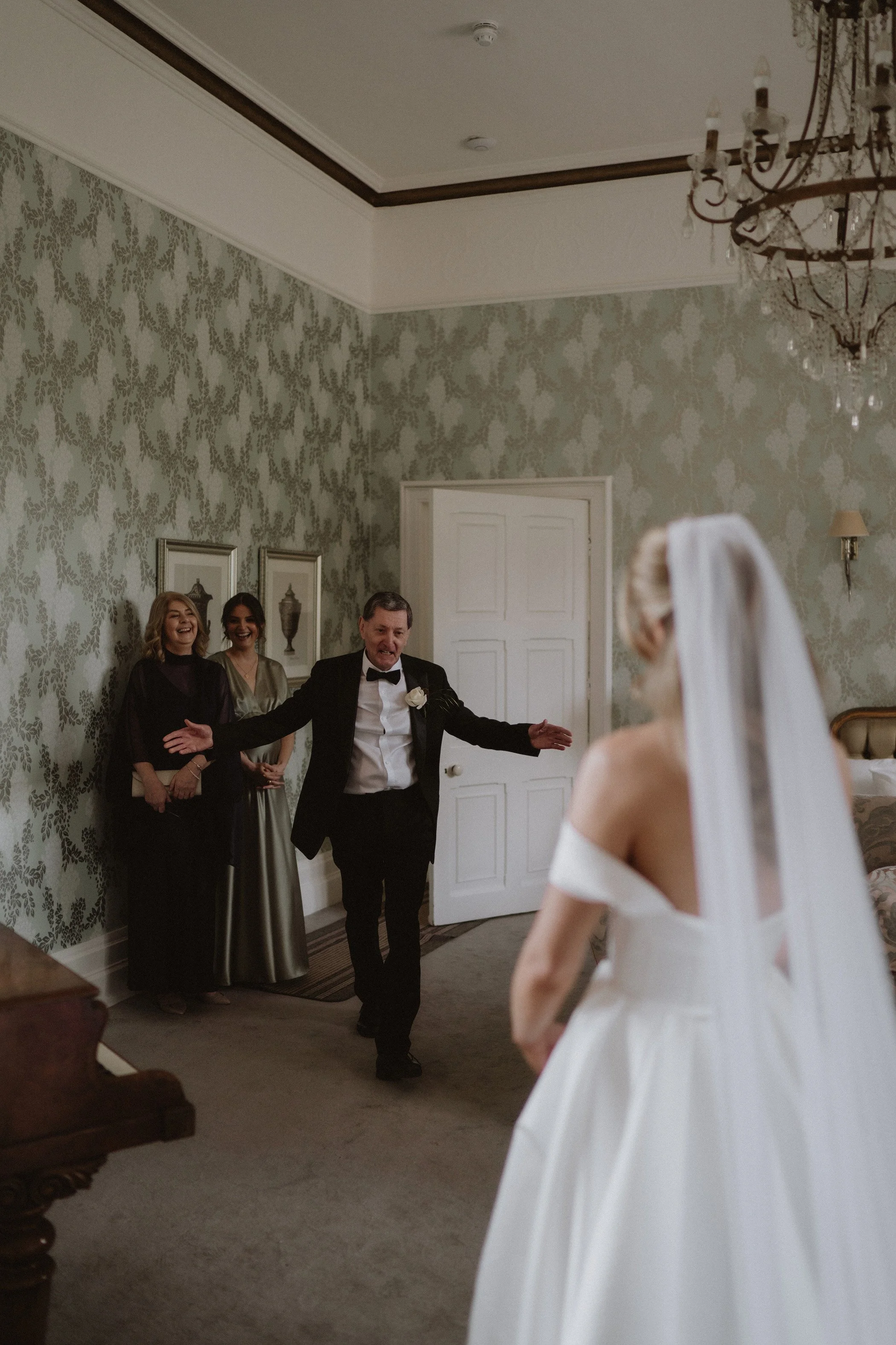 A bride in a white wedding dress and veil facing three people, a man in a tuxedo with a white boutonniere and two women in evening dresses, as the man gestures towards her in a room with ornate wallpaper, chandelier, and closed door.
