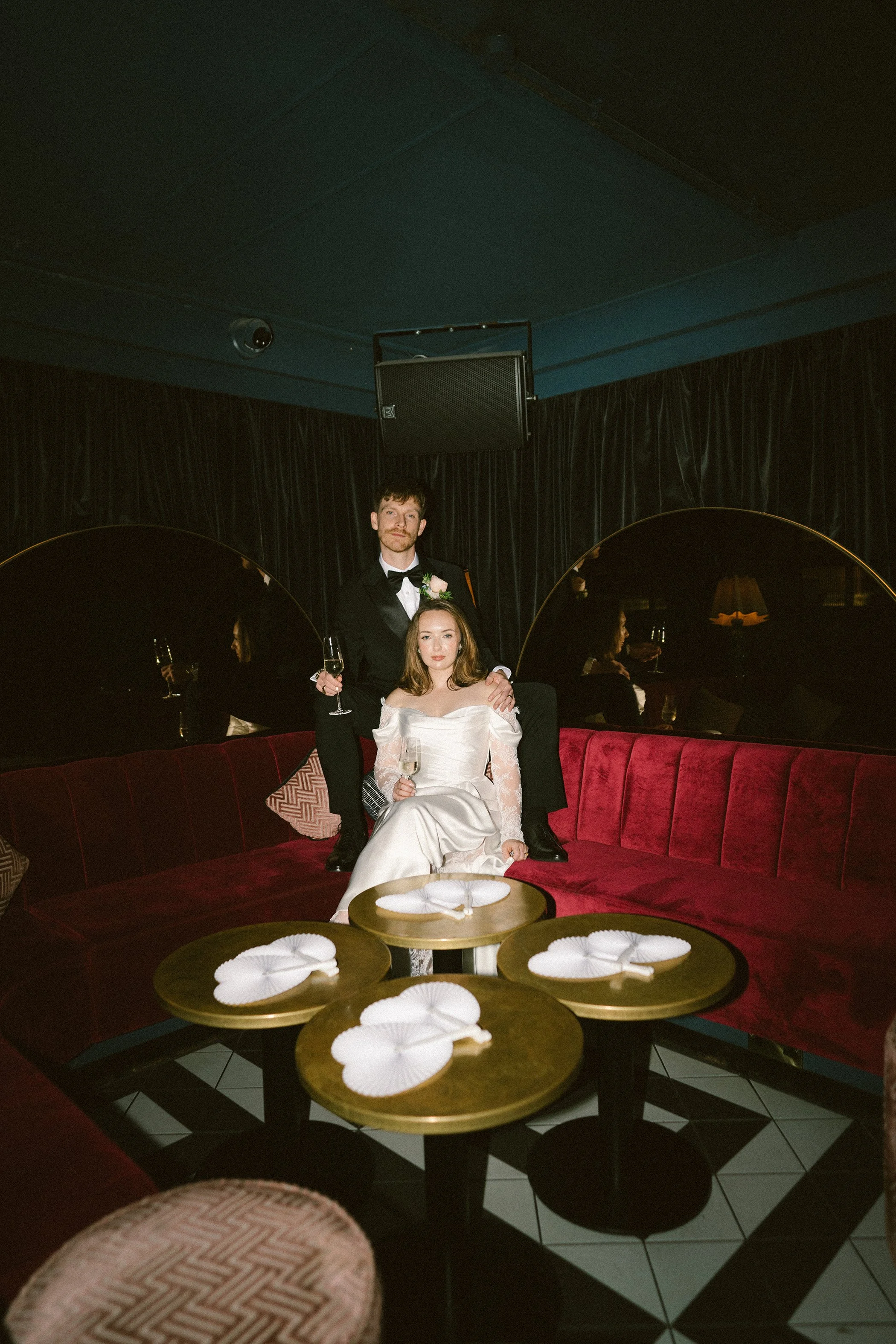A man wearing a tuxedo and a woman wearing a white dress sitting on a red velvet couch in a dimly lit room with black curtains. The man has his arm around the woman, and both are holding glasses of champagne. There are three small round tables with w