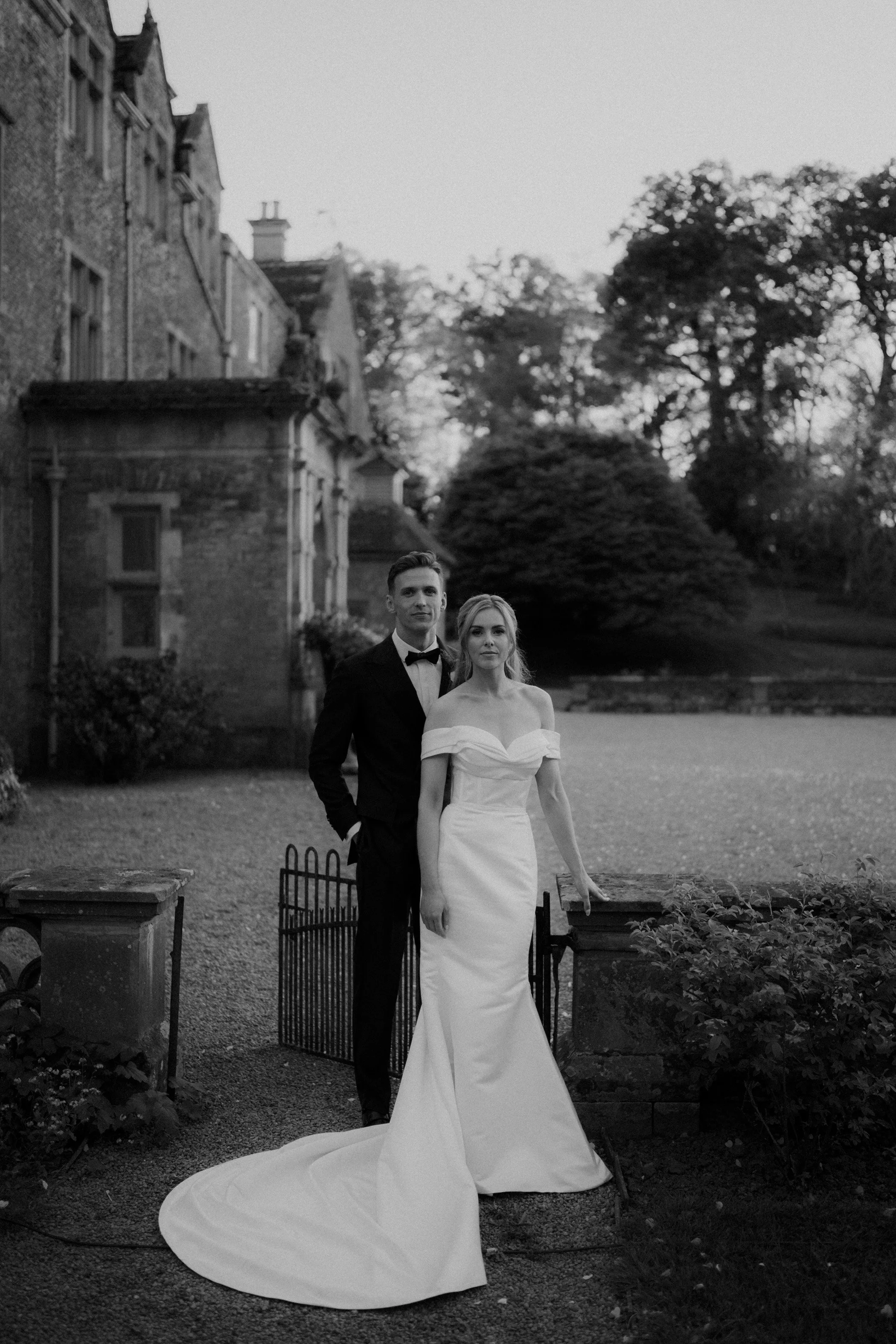 Black and white photo of a bride in a wedding dress and a groom in a tuxedo standing outdoors near a small gate with an elegant house and trees in the background.