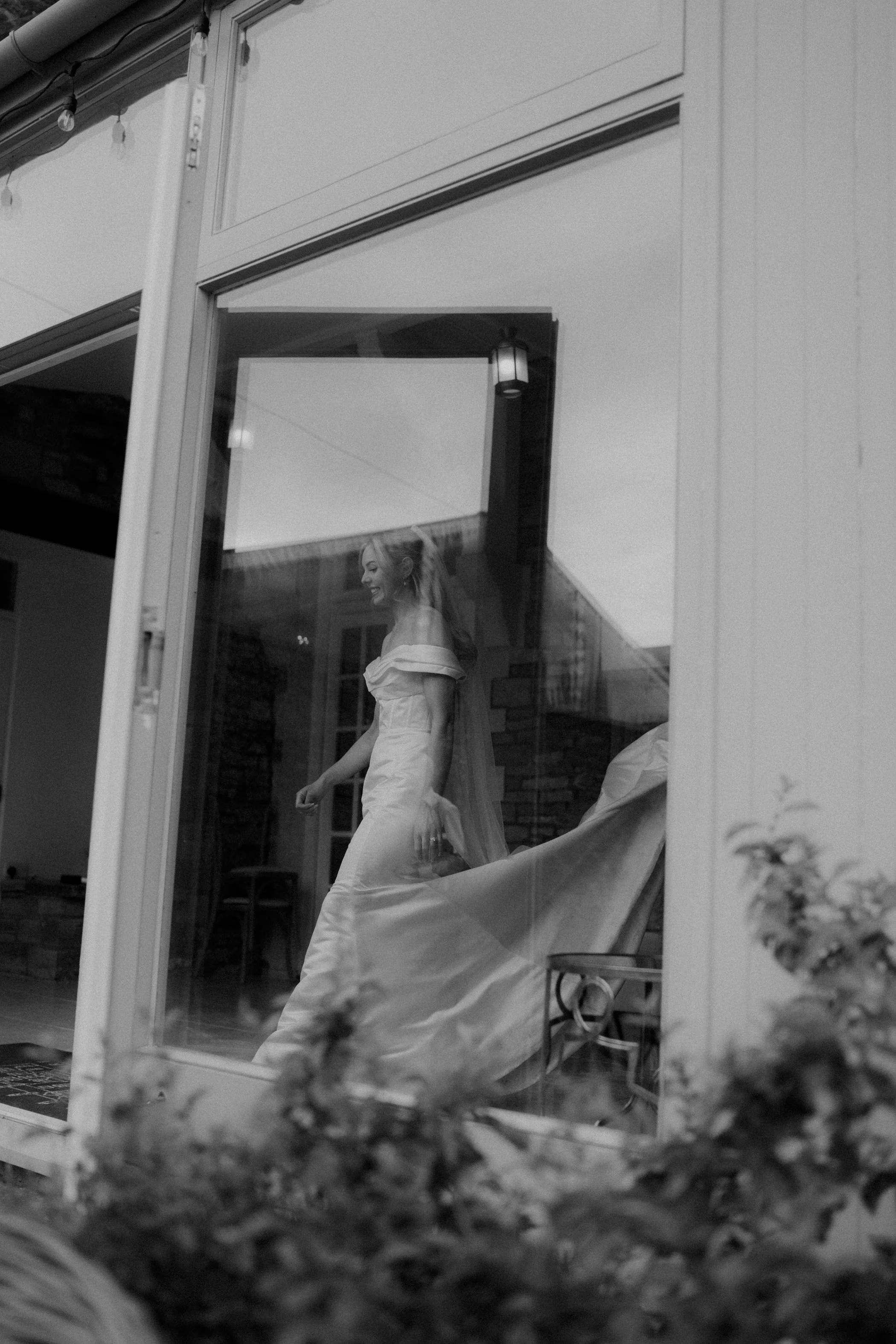 A woman in a wedding dress seen through a glass window, smiling and walking inside a house.