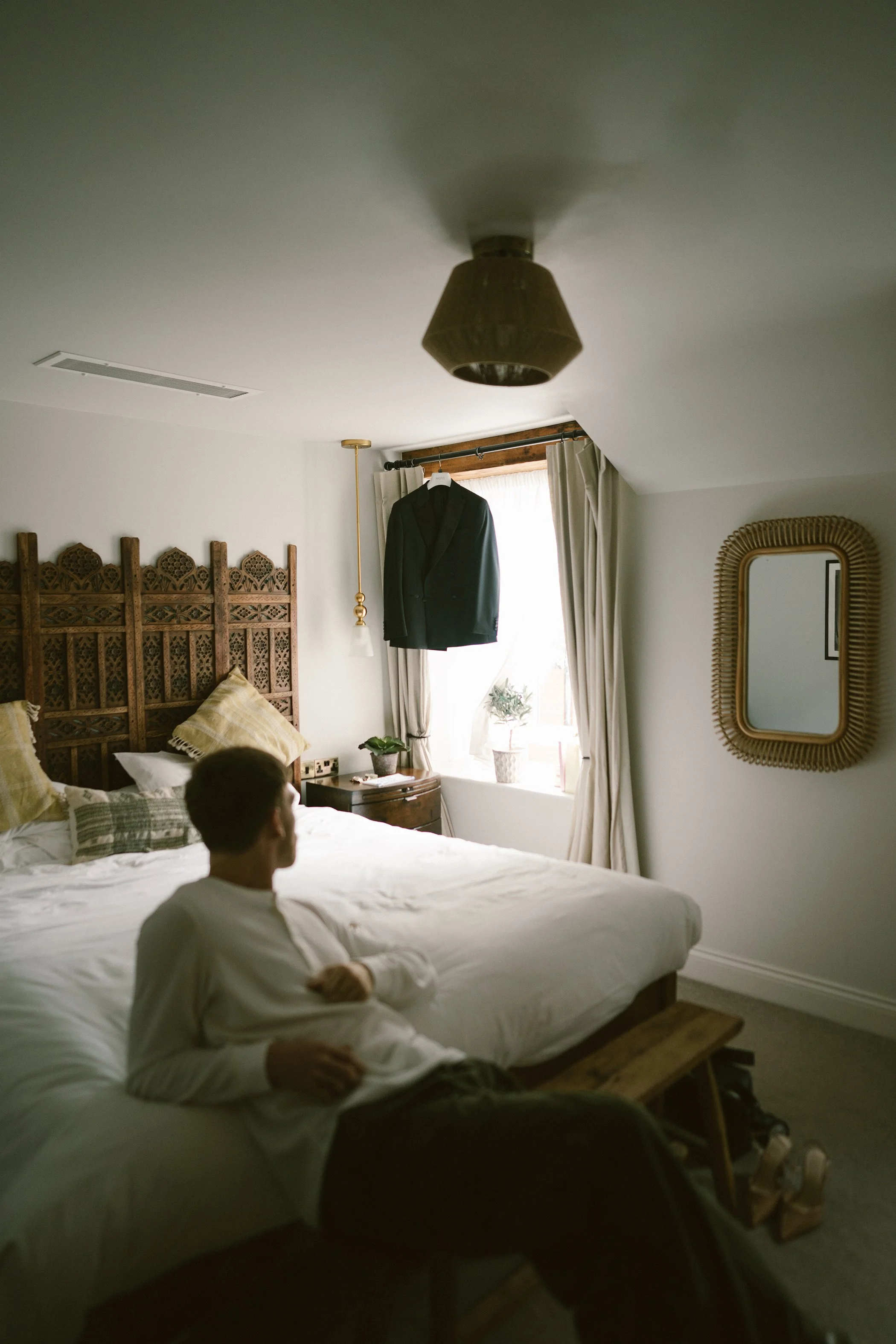 A bedroom with a large bed, a wooden headboard, and a person sitting on the bed looking to the side. There is a window with white curtains, a black suit jacket hanging in front of it, and a mirror on the wall. The ceiling has a light fixture, and the