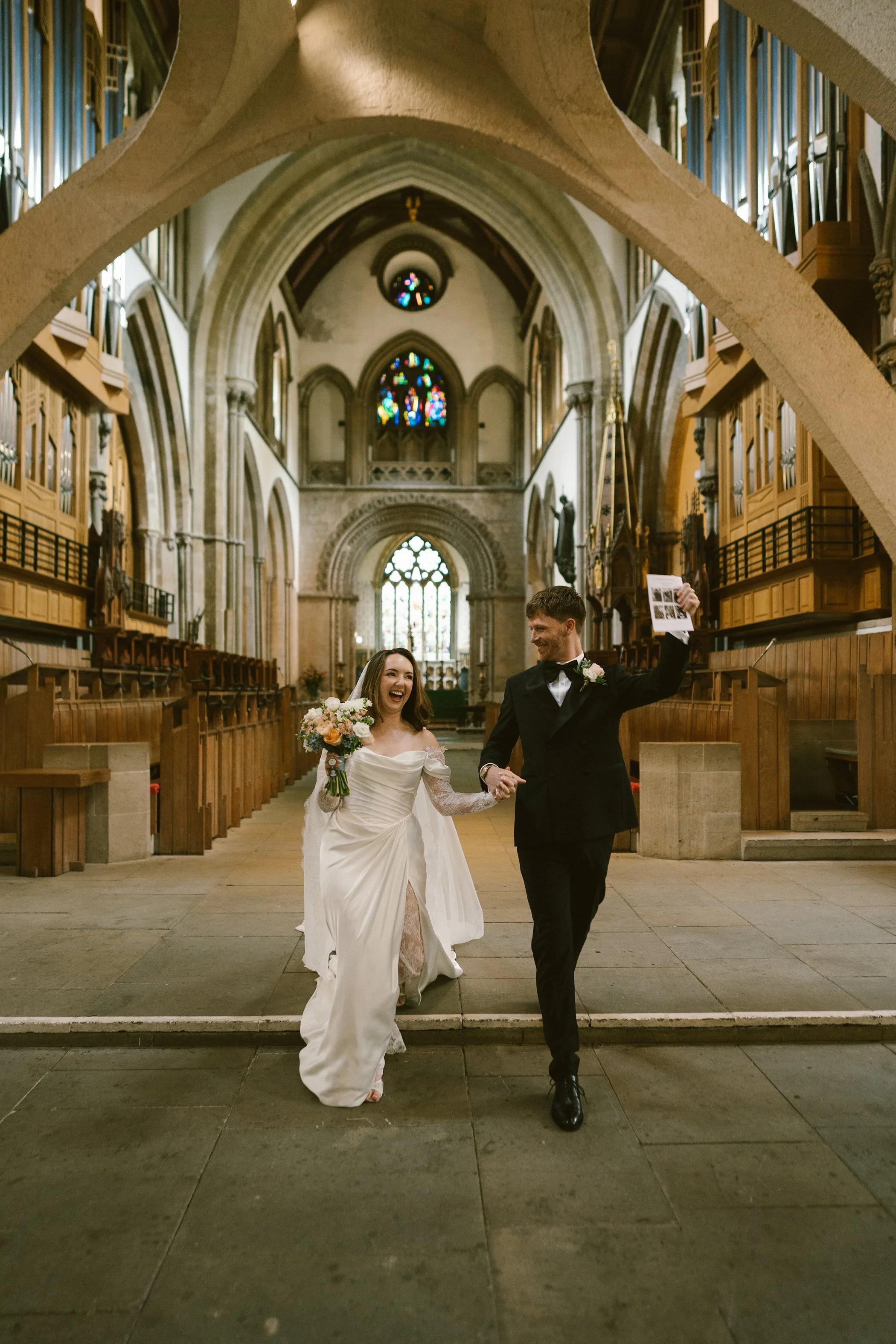 A newly married couple walking inside a church. The bride is wearing a wedding gown and holding a bouquet, smiling happily. The groom is wearing a tuxedo and appears to be celebrating, holding up a photo. The church has stained glass windows and orna
