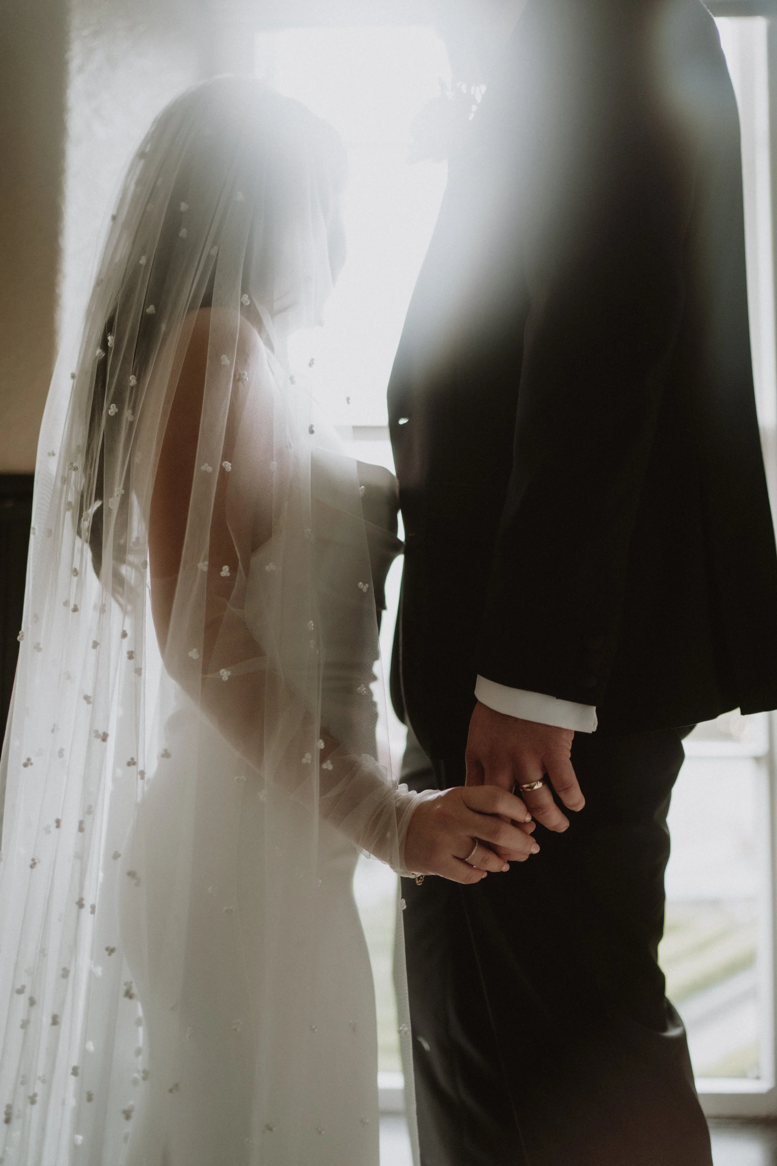 Bride and groom holding hands during their wedding ceremony, with the bride wearing a veil and wedding dress and the groom dressed in a suit, standing near a window with bright light shining through.