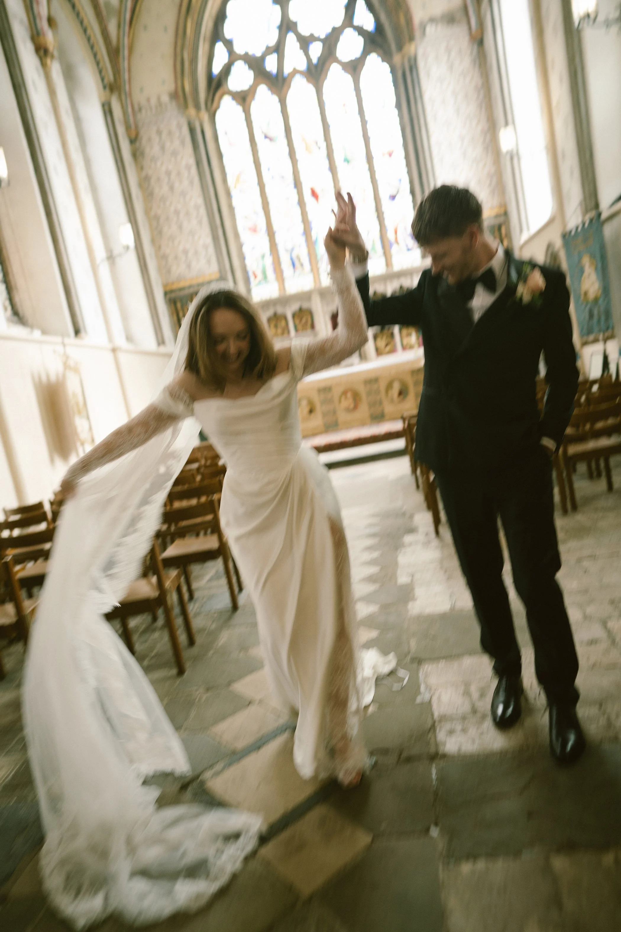 A bride and groom dancing inside a church with wooden pews and stained glass windows. The bride is wearing a white wedding dress and veil, and the groom is in a black tuxedo. They appear to be joyful, holding hands and smiling.
