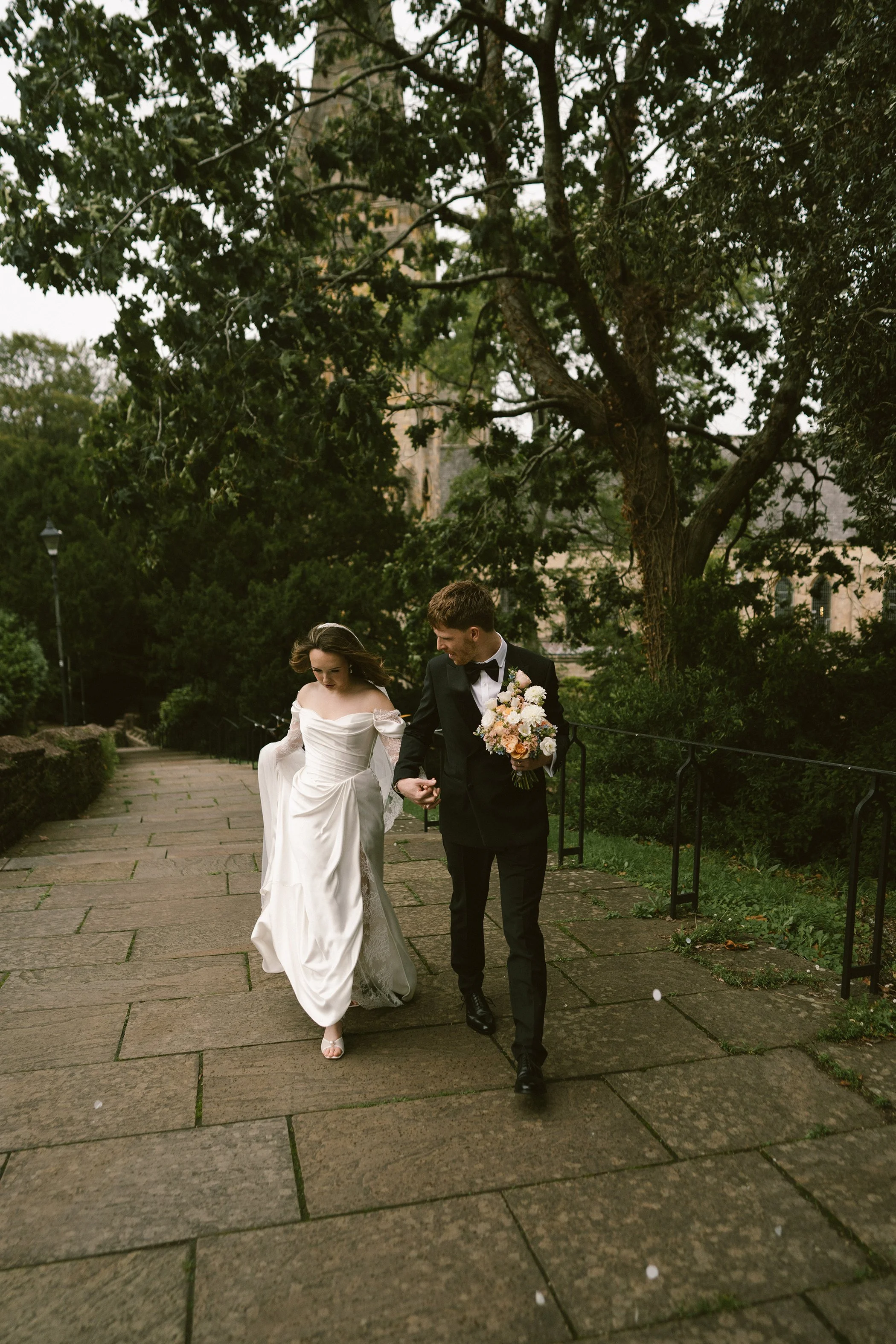 A bride in a white wedding gown holding a groom's hand in a black tuxedo, walking together outdoors on a paved path surrounded by greenery and trees.