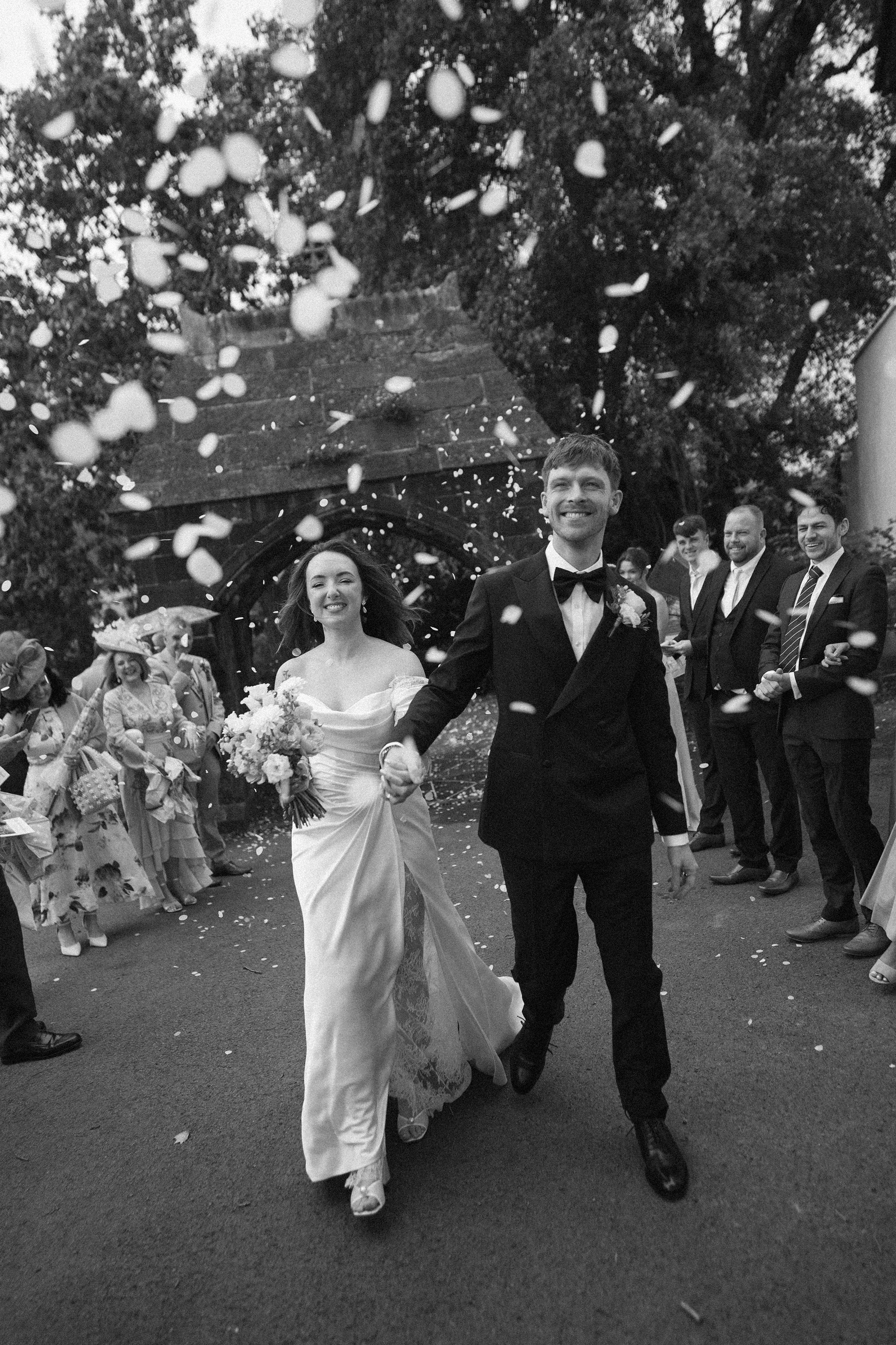 Black and white photo of a wedding celebration with a bride and a groom walking hand in hand among guests outside. The bride wears a long white dress and carries a bouquet, smiling. The groom wears a black tuxedo with a bow tie, smiling. Guests are o