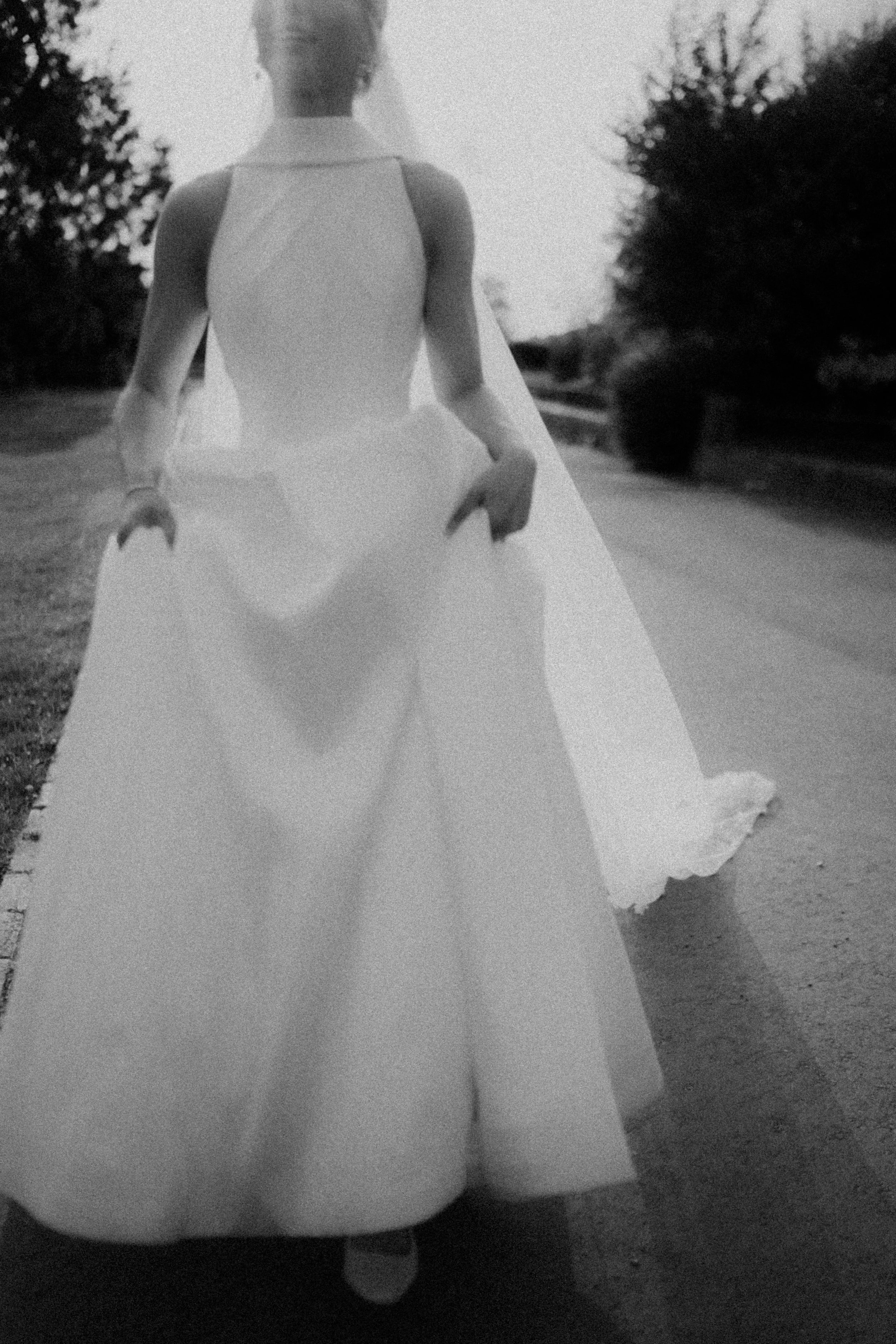 A woman in a long, elegant white gown with a cape walks on a paved path. The photo is in black and white, with trees in the background. south wales uk wedding