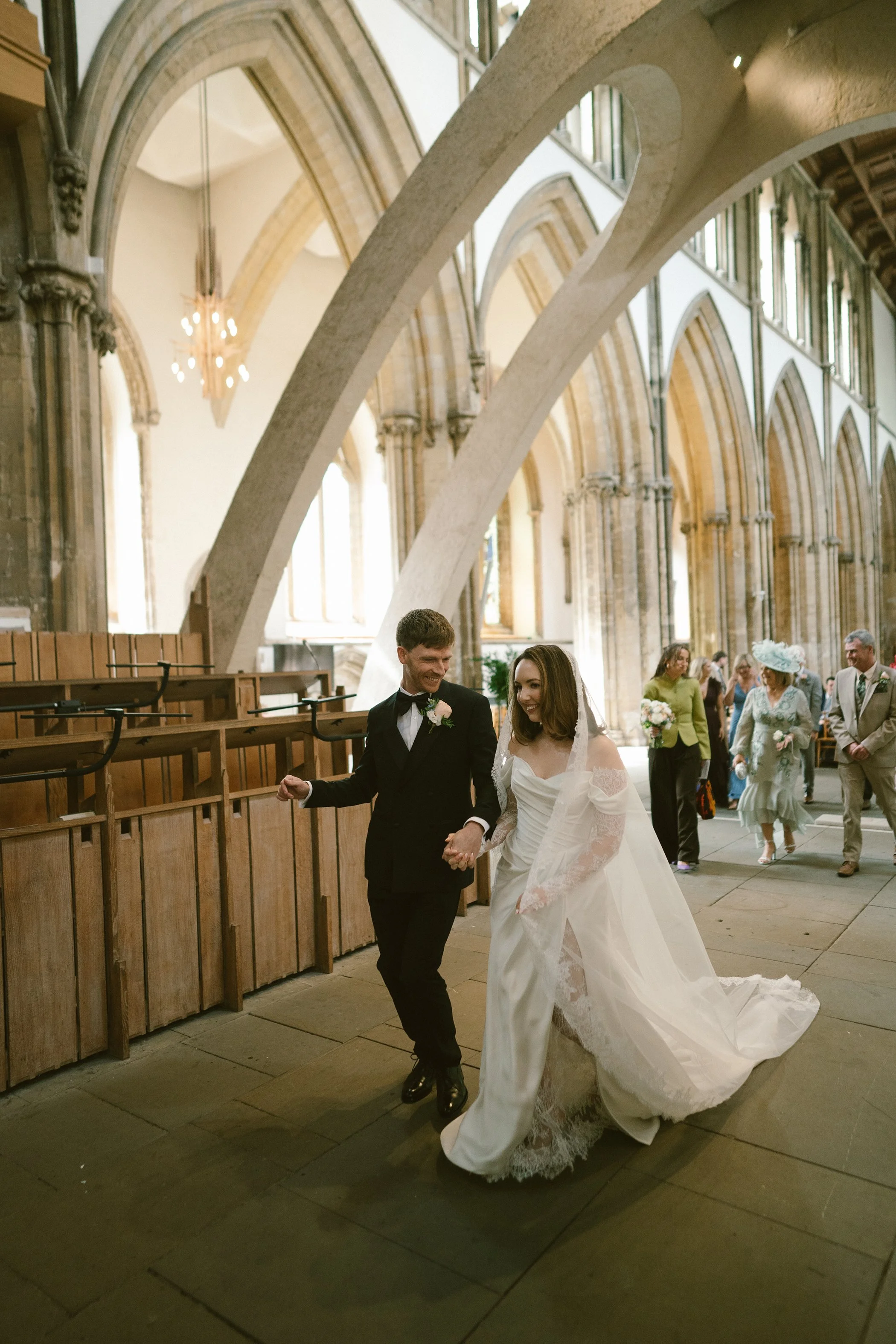 A bride and groom walking hand in hand inside a church after their wedding ceremony, with guests in the background.