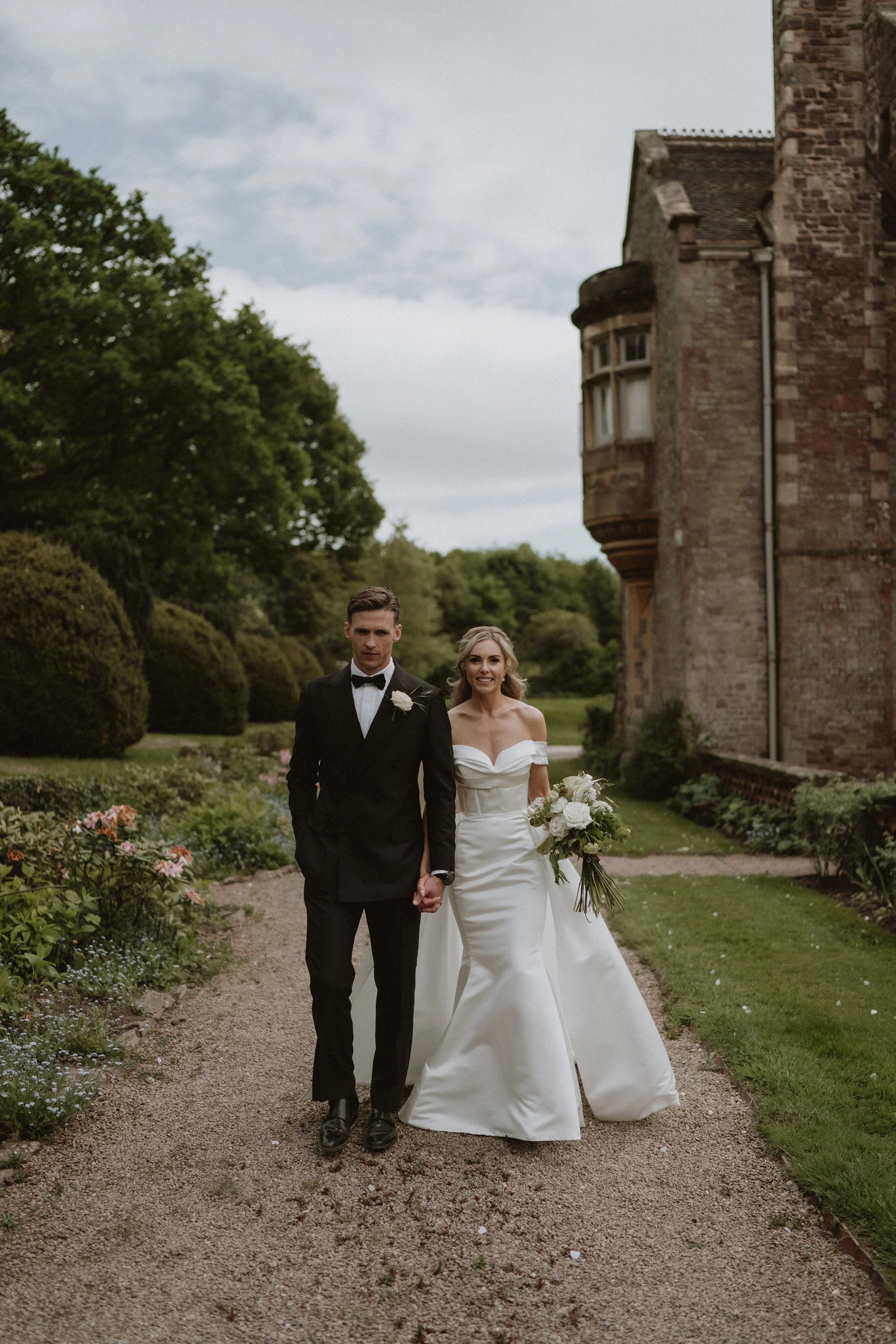 A bride and groom walking hand in hand on a garden path during their wedding day, with a historic building in the background.