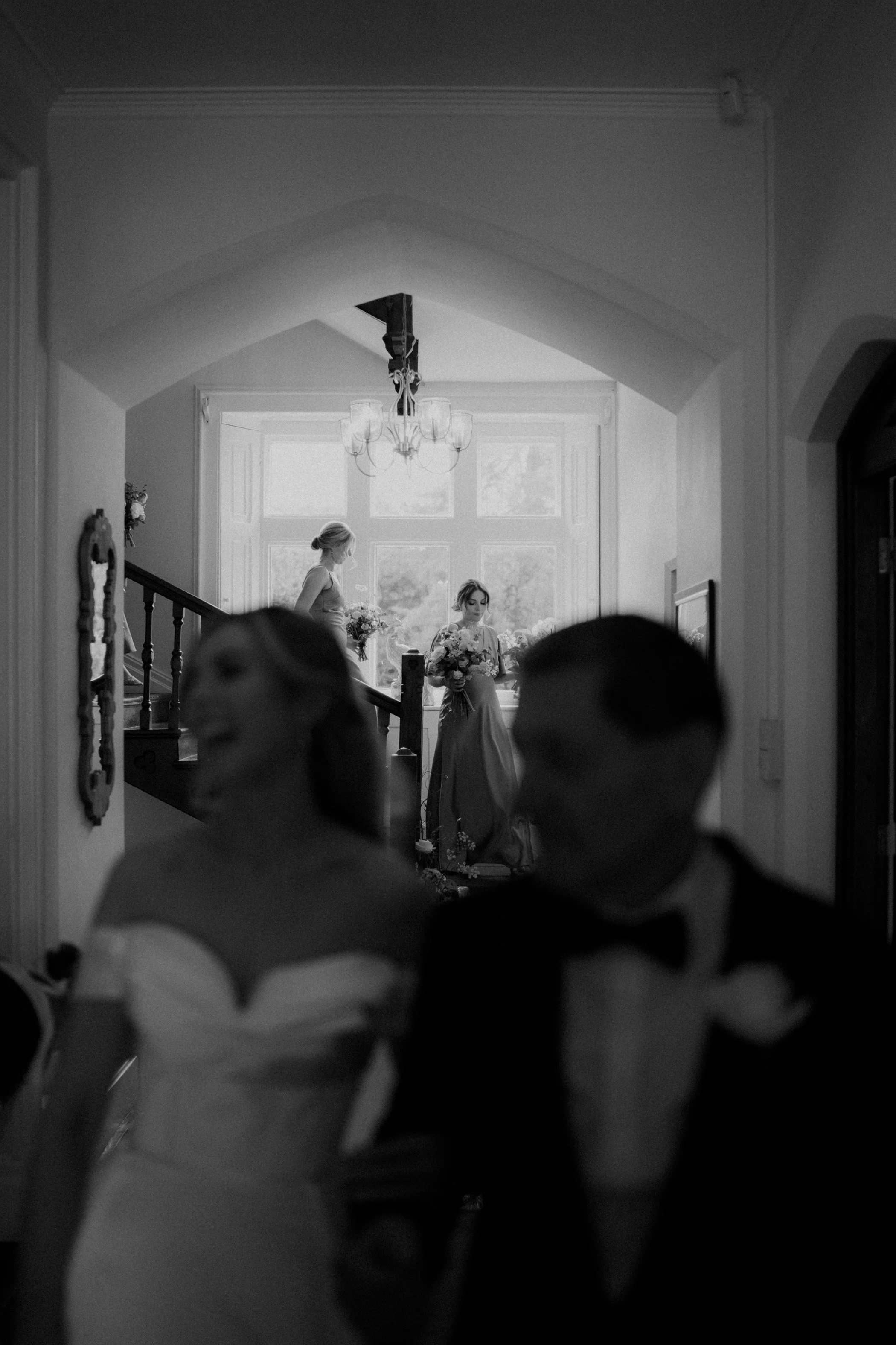 Black and white photo of a wedding scene with a bride and groom in the foreground, blurred, and two women with bouquets standing near a staircase in a bright room with large windows in the background.