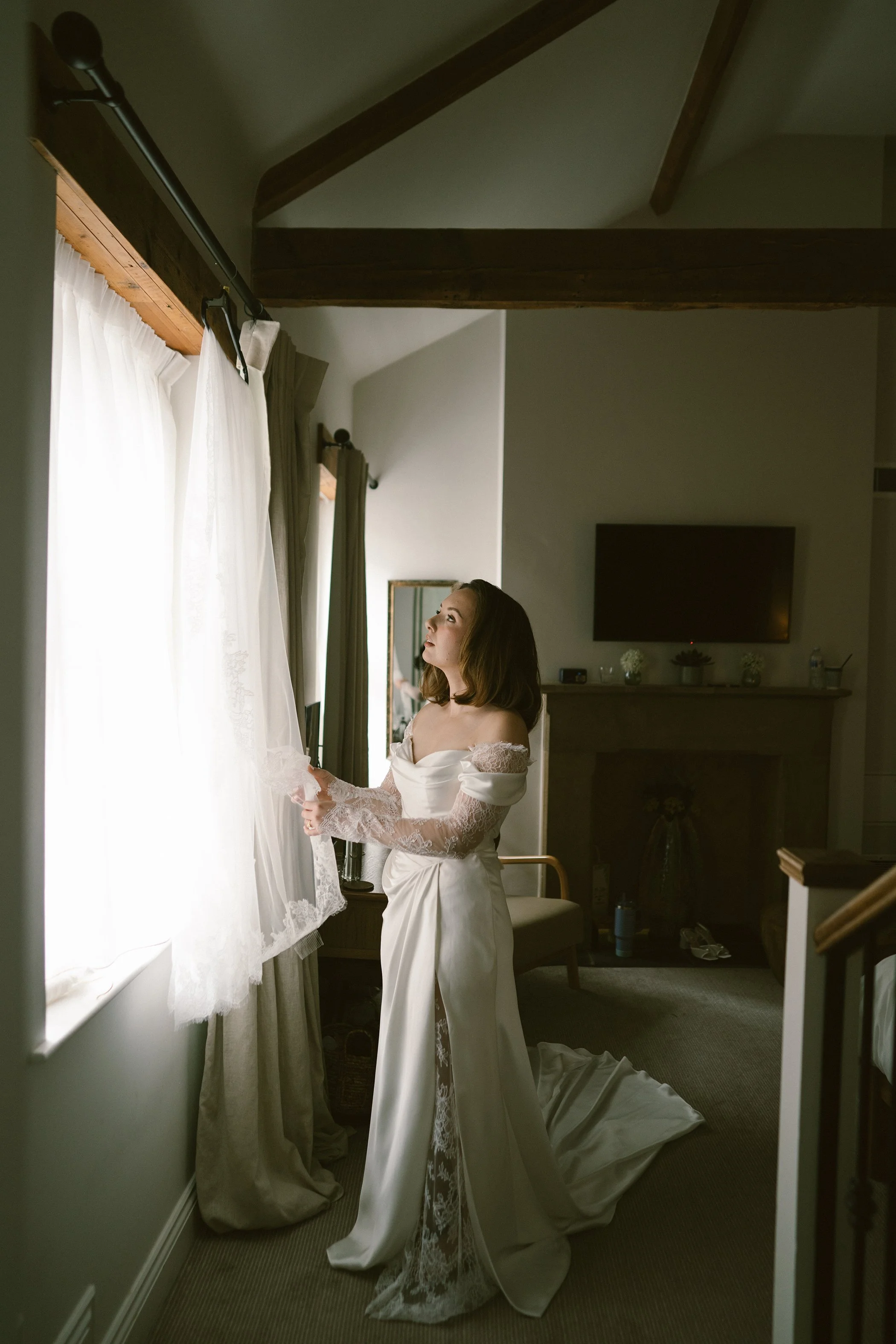 A woman in a satin wedding gown with lace details on the sleeves stands by a window, holding and looking at white curtains, with natural light illuminating her face in a cozy room.