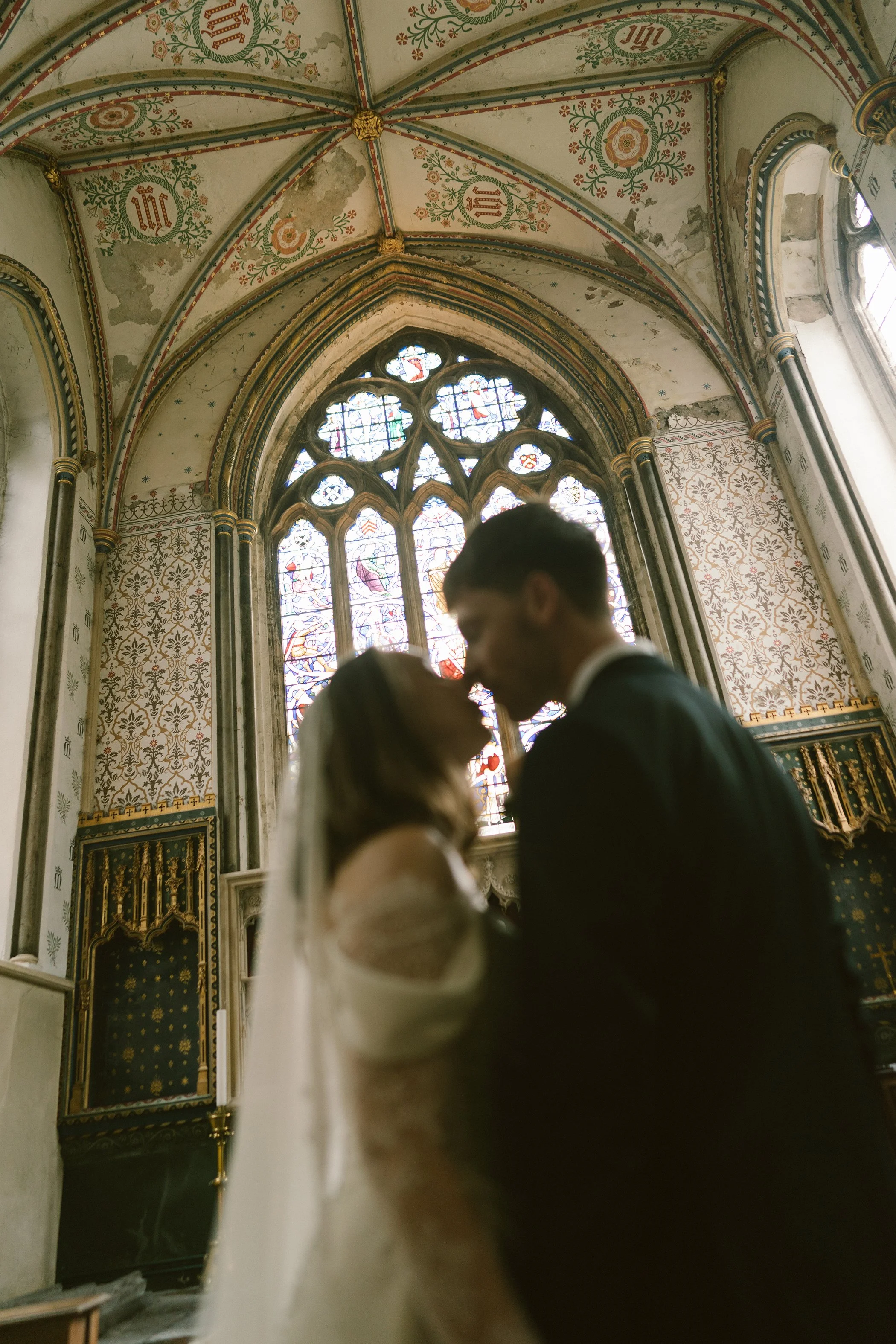A couple dressed in wedding attire sharing a kiss inside a historic church with ornate stained glass windows, intricate painted ceiling, and decorative walls.