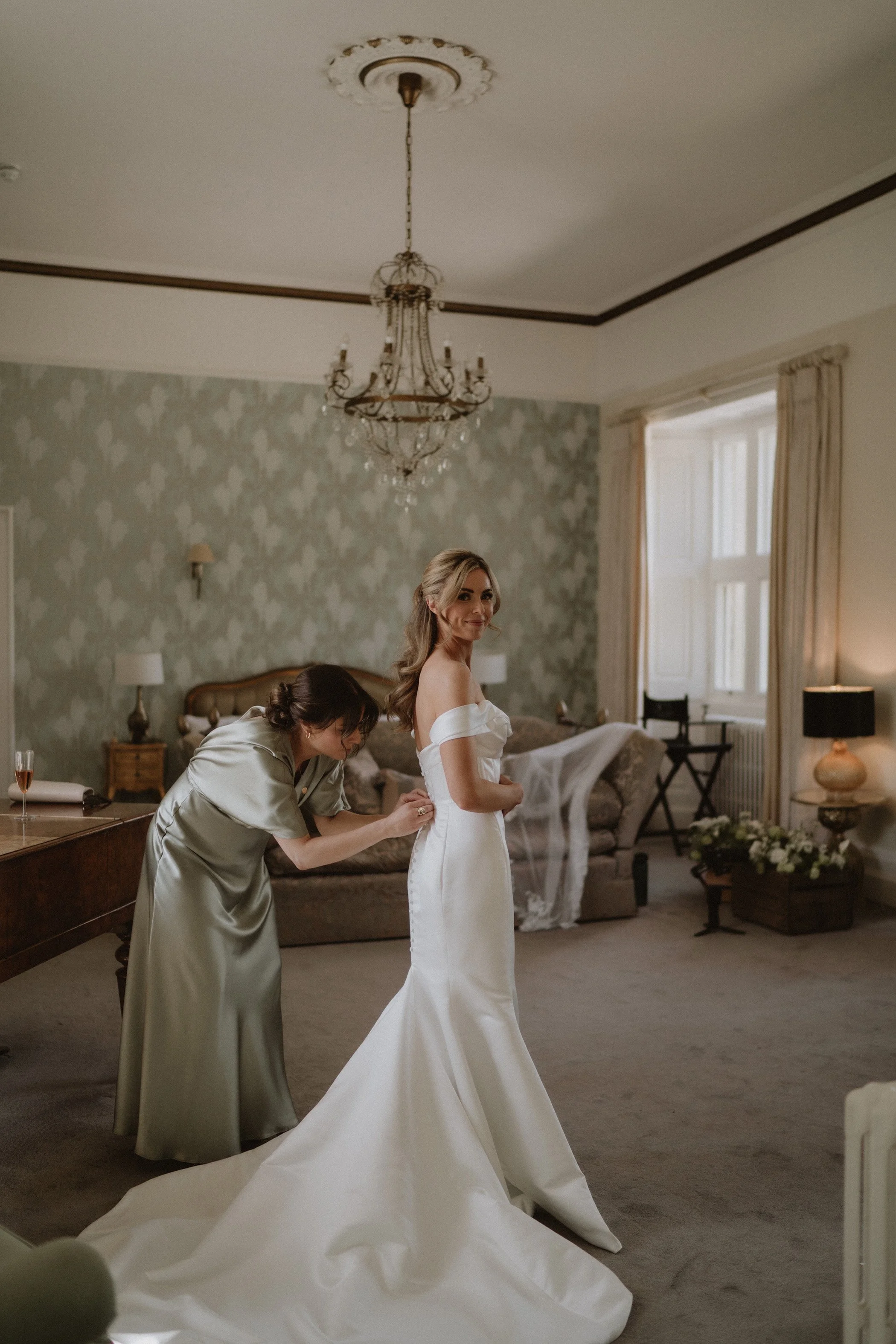 A bride in a white off-shoulder wedding dress is getting ready in a vintage-style room with a woman helping her with the dress.