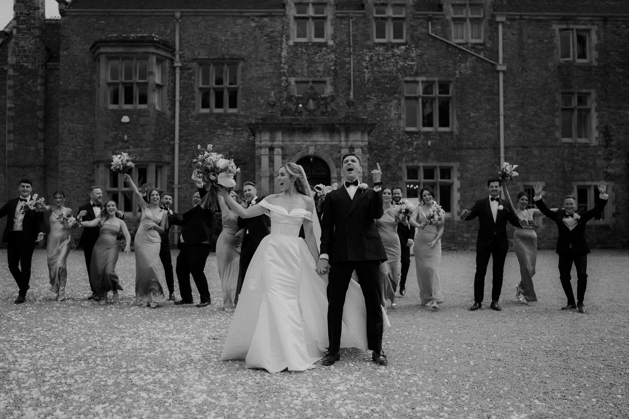 A black and white photo of a wedding party standing outside a large old building, with the bride and groom at the front holding hands, and the bridesmaids and groomsmen celebrating in the background.