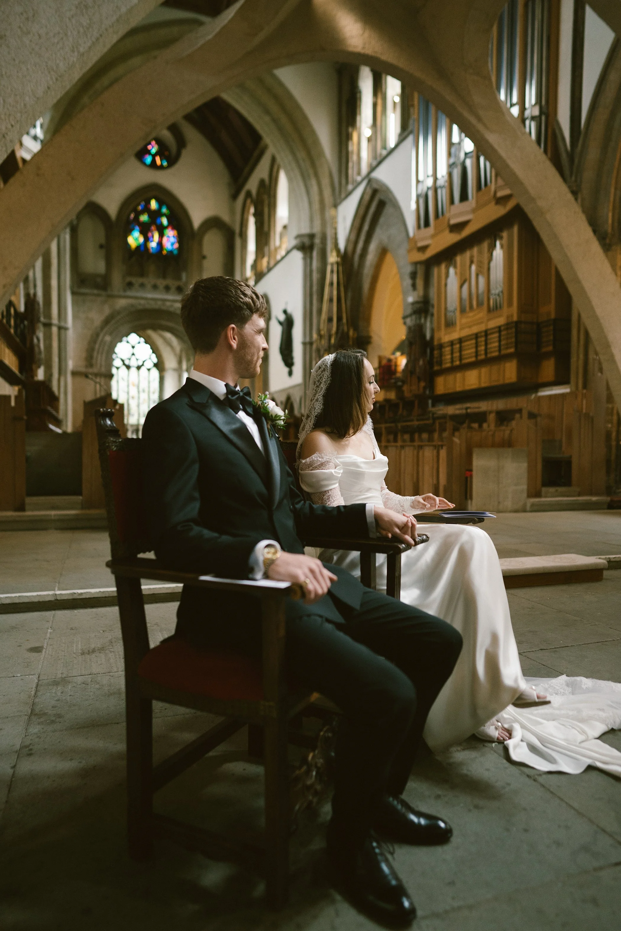 A bride and groom sit side by side in a church with tall stained glass windows, wearing formal wedding attire, with their heads bowed.
