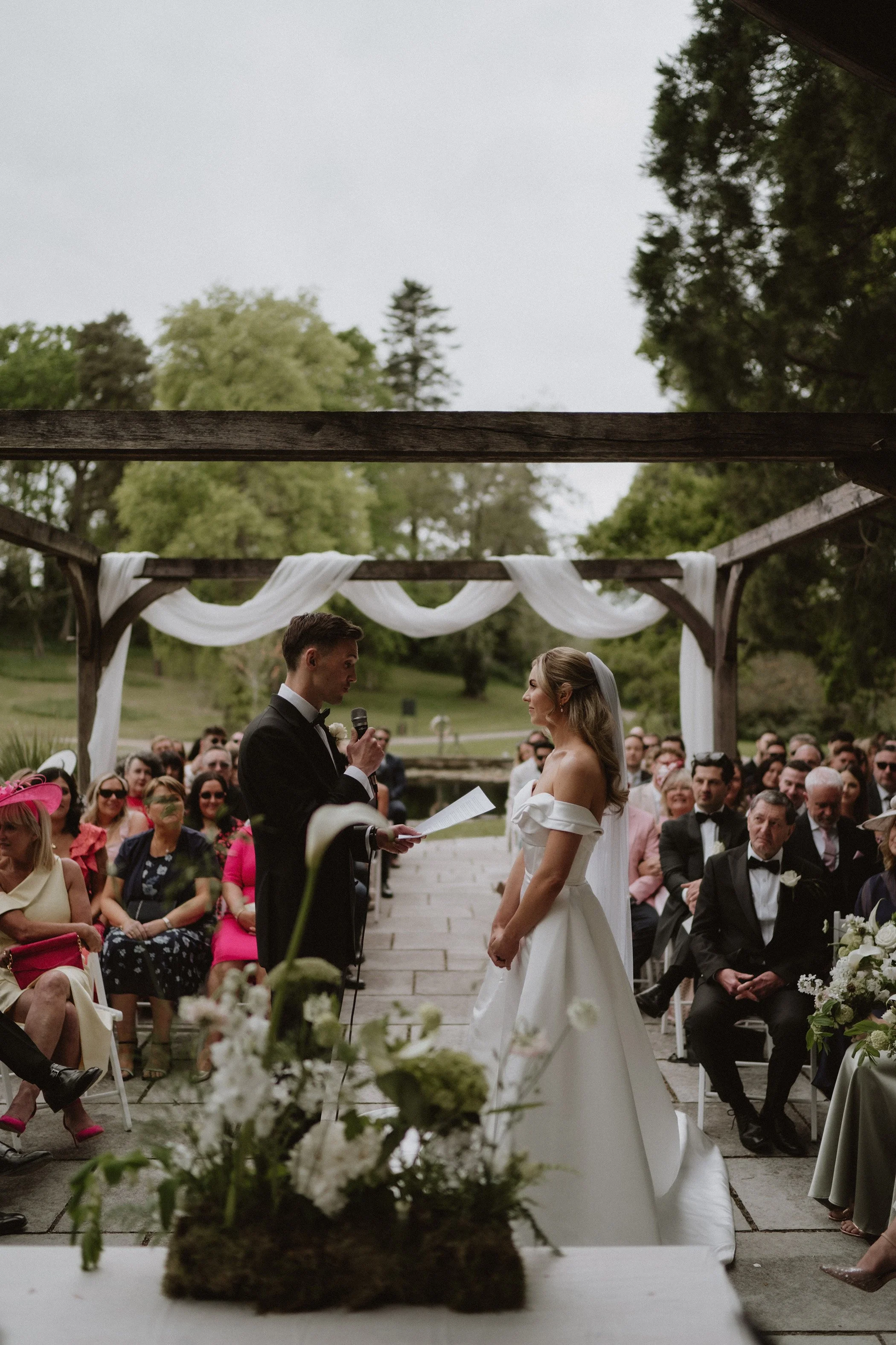 A bride and groom standing face-to-face during a wedding ceremony outdoors, with guests seated around them under a decorated wooden pergola, green trees in the background.