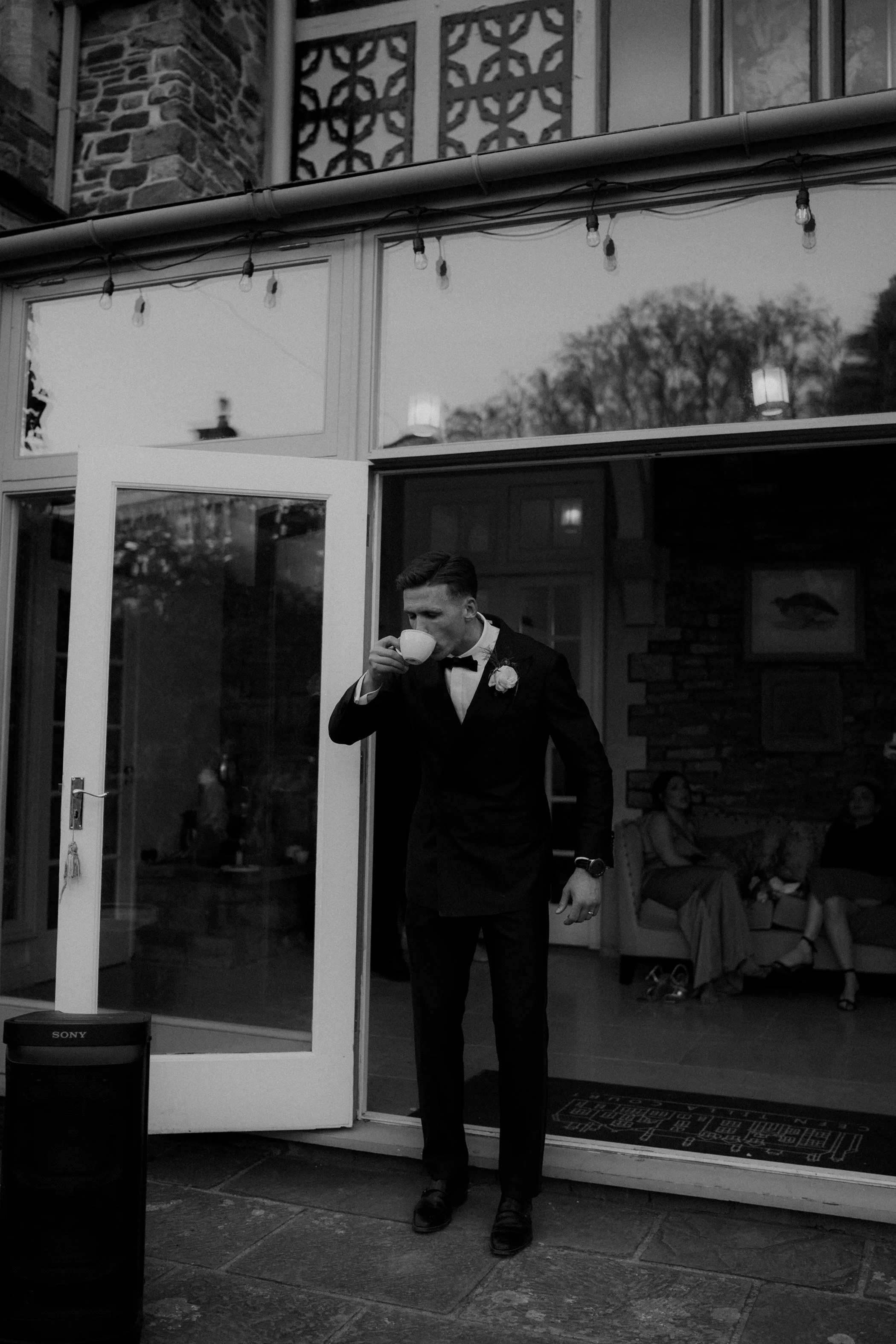 A young man in formal attire, including a bow tie and boutonniere, drinking from a cup in front of a house with string lights and large windows, with three women sitting inside.